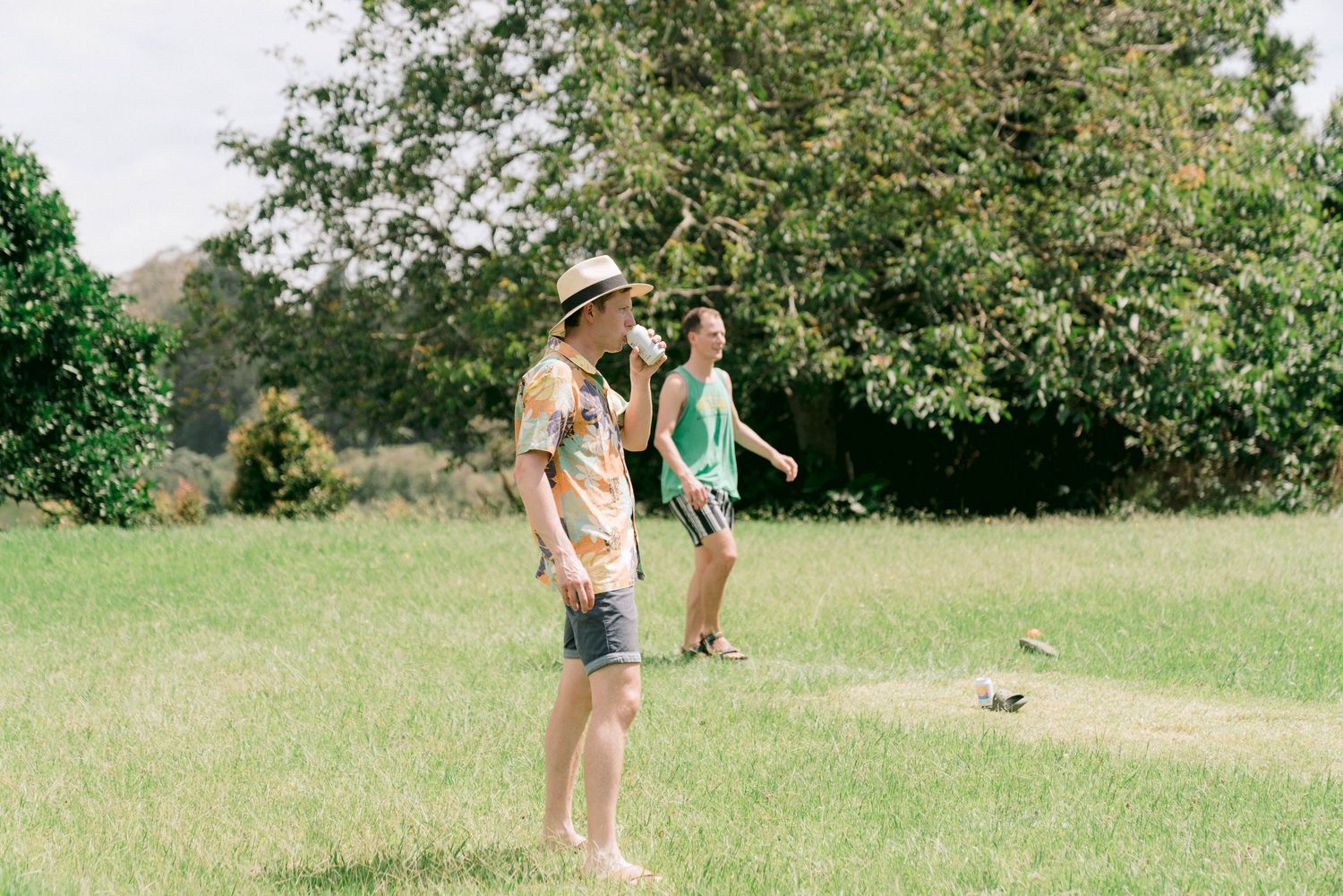 A couple enjoys a sunny day outdoors with a small dog in a grassy field surrounded by trees.