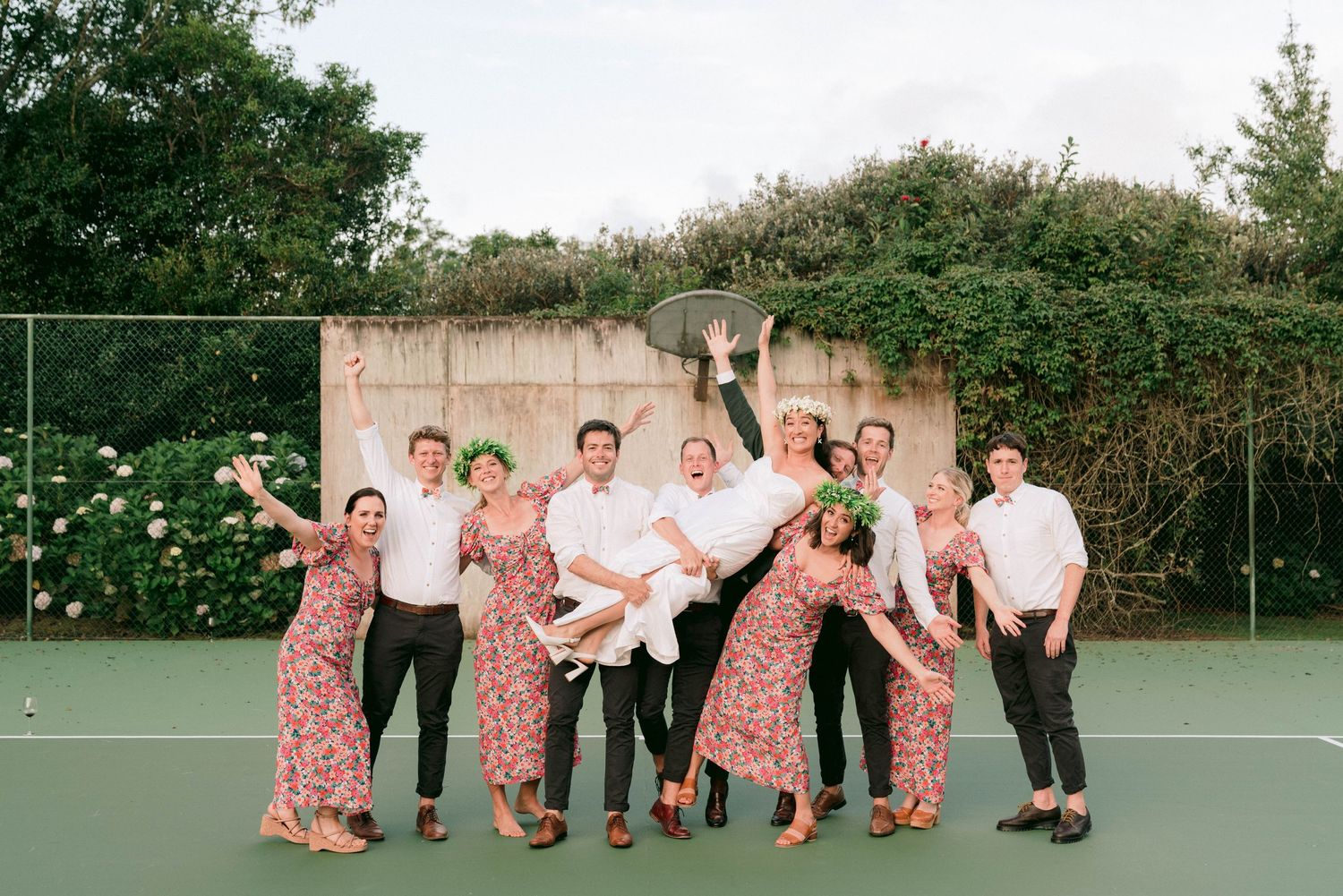 Wedding party poses playfully on a tennis court with bridesmaids wearing matching floral dresses.