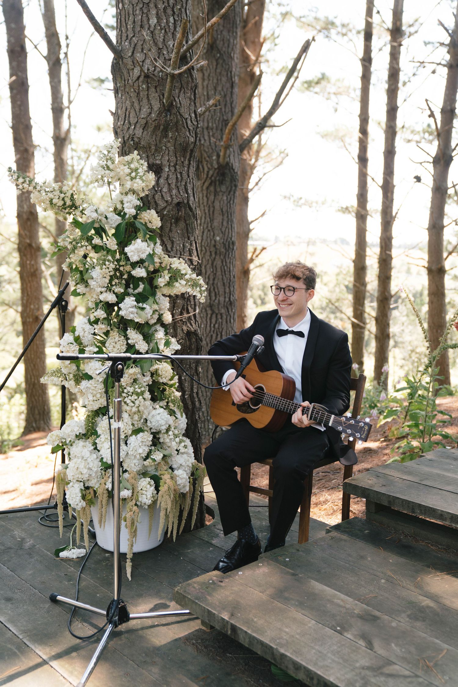A guitarist in a tuxedo performs next to elegant white floral arrangements in a wooded outdoor wedding setting.
