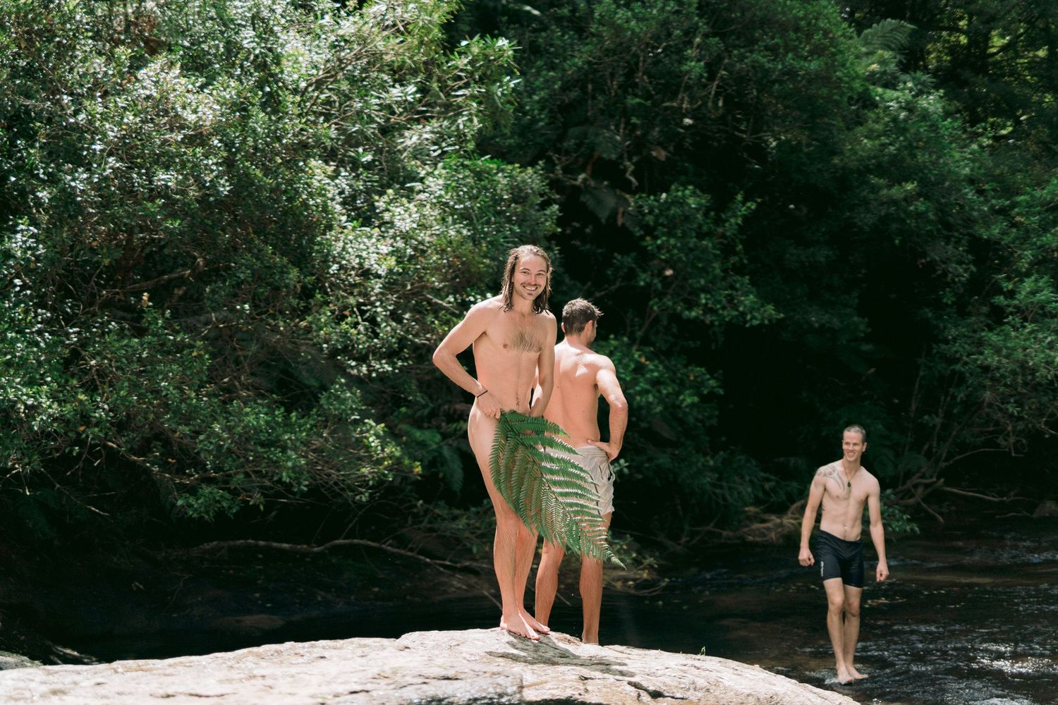 People enjoying a summer day standing on rocks by a creek in a wooded area.