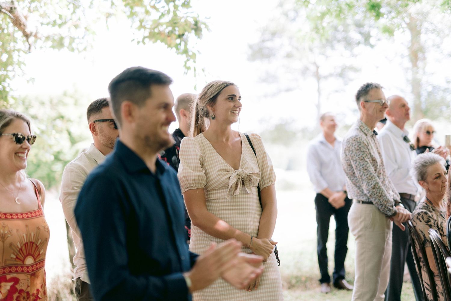 Wedding guests standing together during an outdoor ceremony beneath trees.