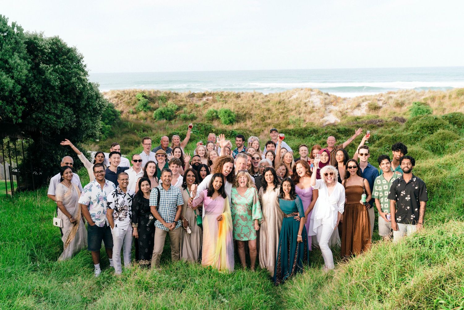 Large wedding party gathered on a grassy coastal hillside wearing pastel colors.