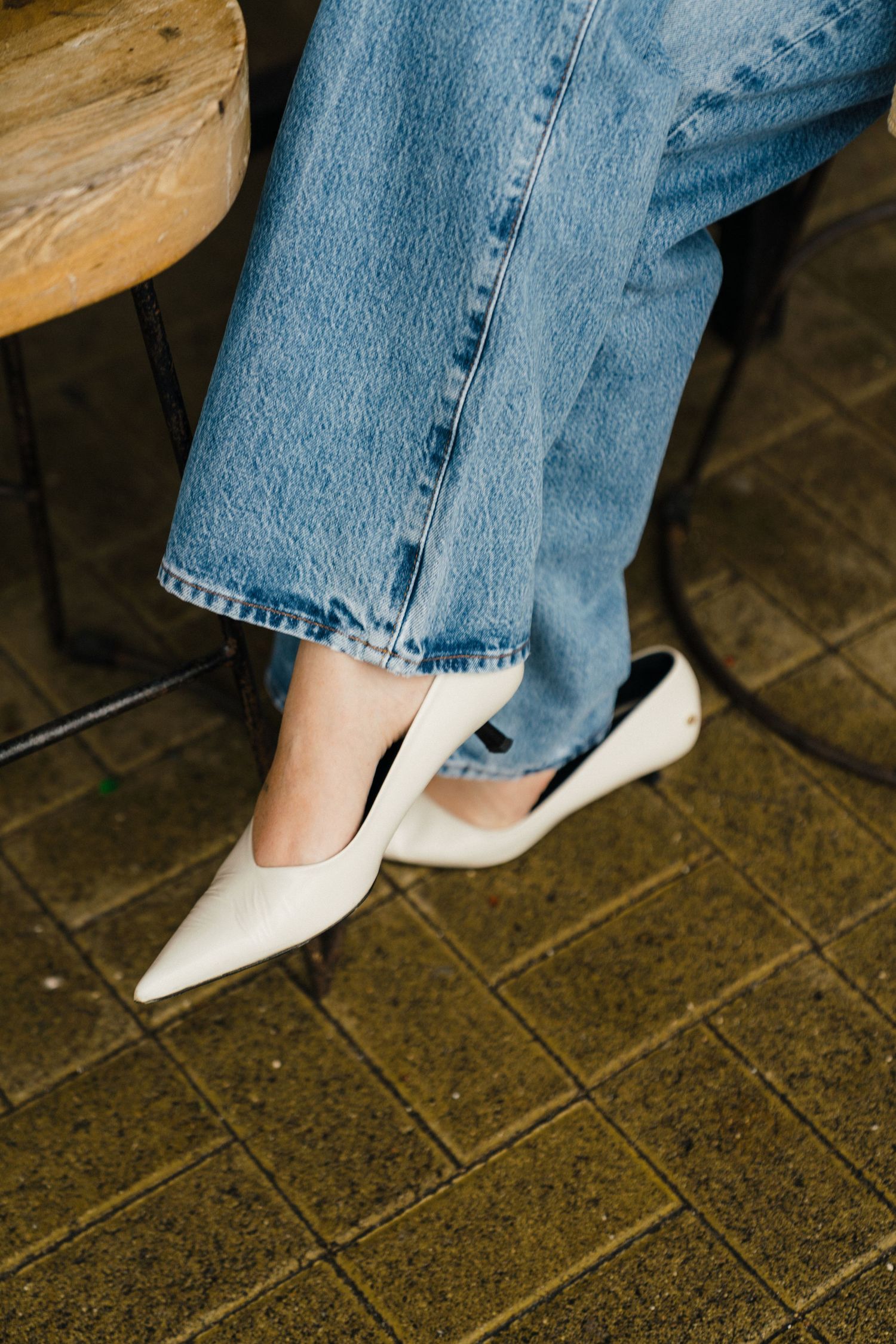 Person wearing classic white pointed ballet flats and light wash denim jeans sitting on a tiled floor.