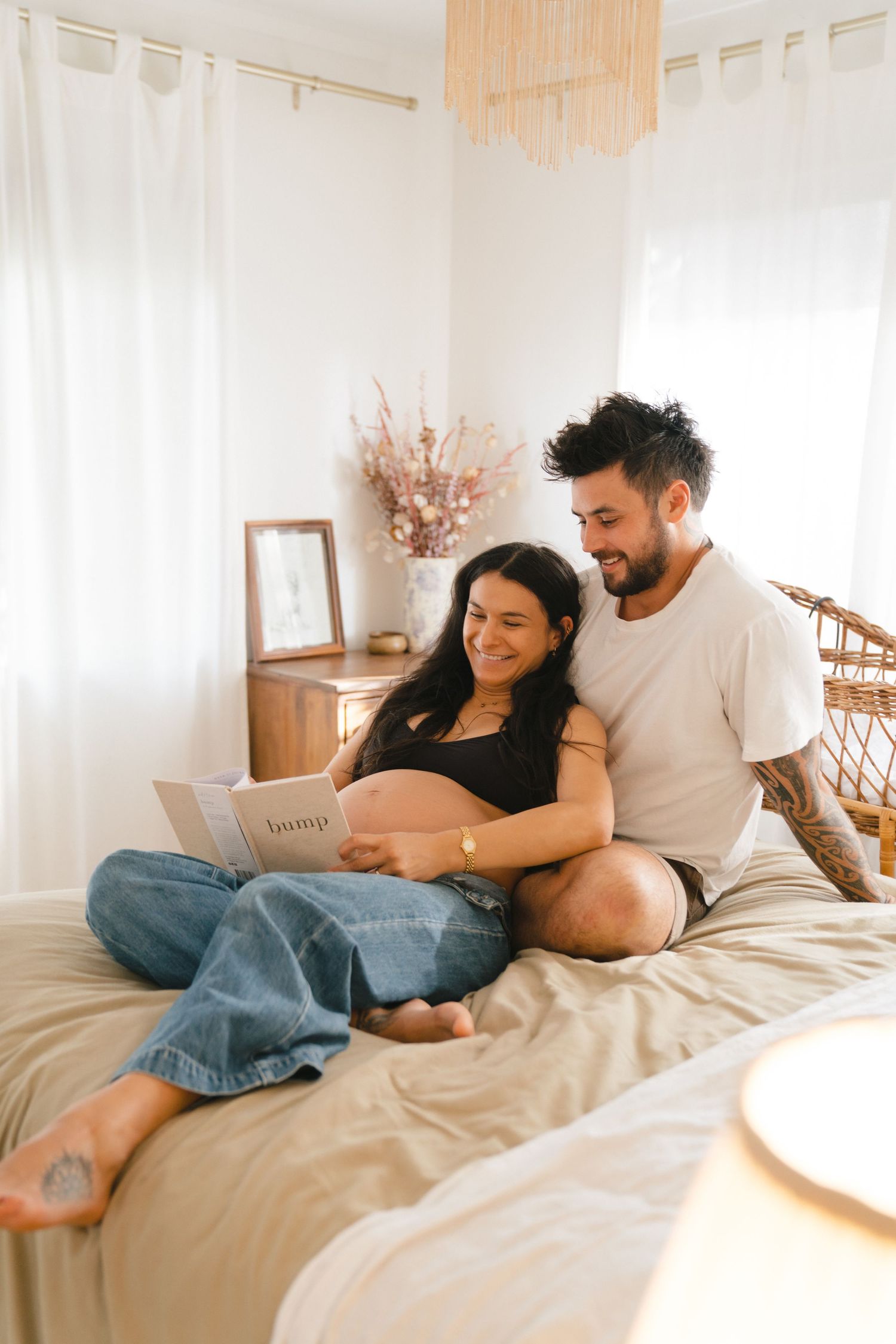 A couple relaxes together on a bed while reading a book in a bright, cozy bedroom with white curtains and decor.