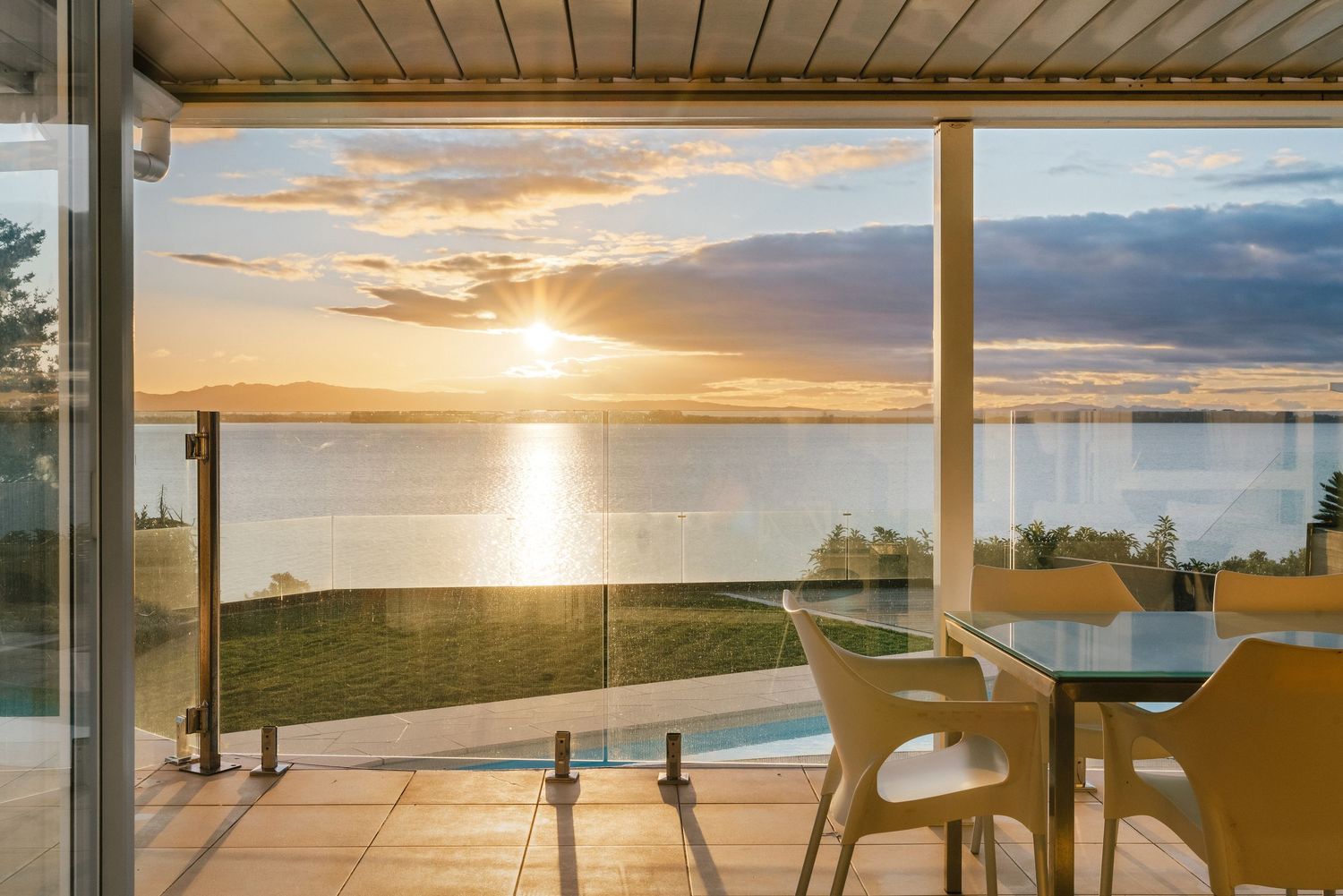 Modern patio dining area with glass railing overlooking waterfront sunset view.