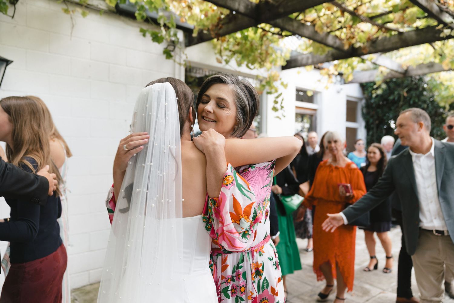 Wedding reception guests mingling under a covered outdoor patio with greenery.