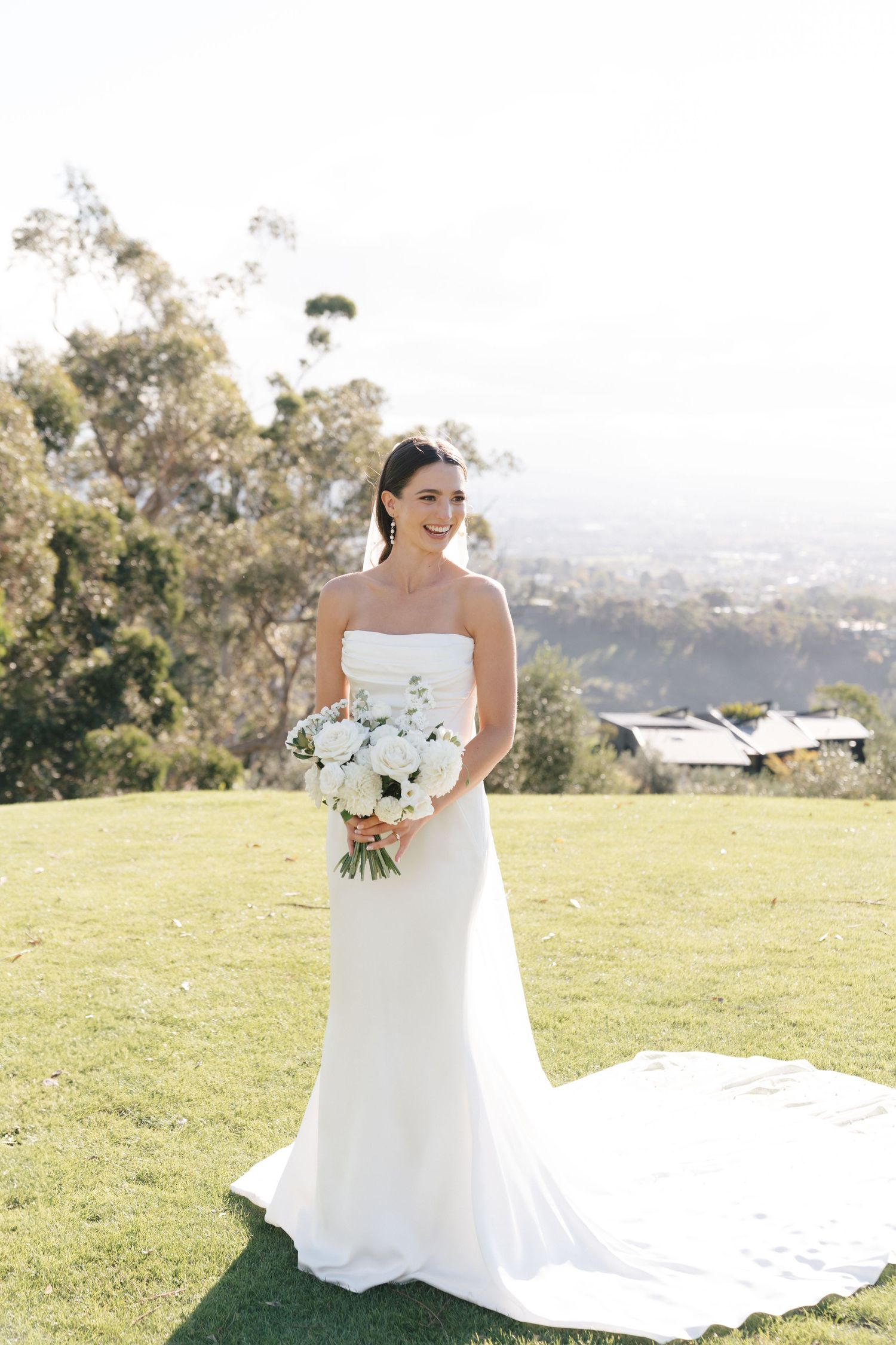 A bride in a white strapless wedding gown holds a white bouquet while standing on a hilltop overlooking a scenic view.