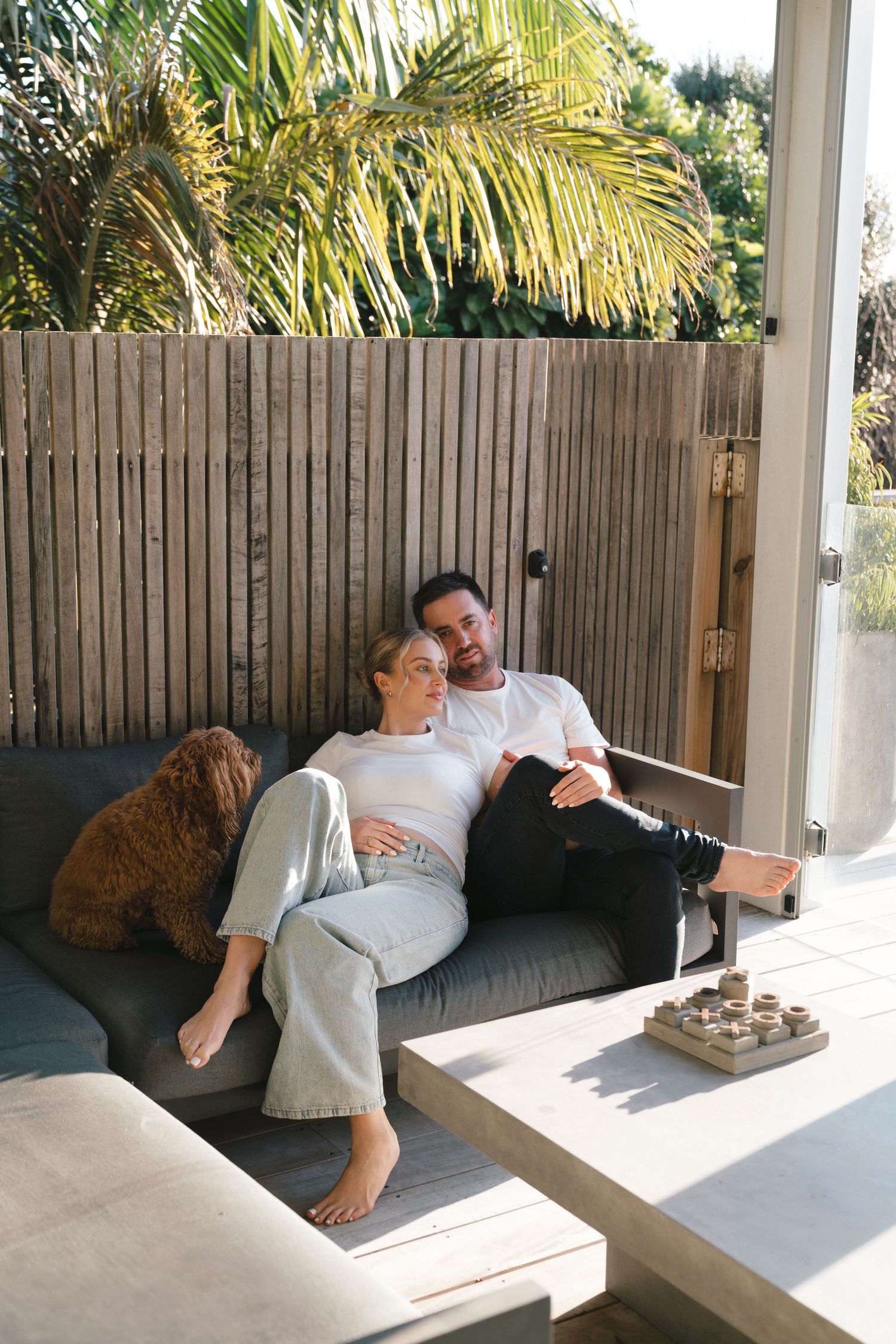 Couple relaxing on outdoor patio couch with palm trees and wooden fence in background.