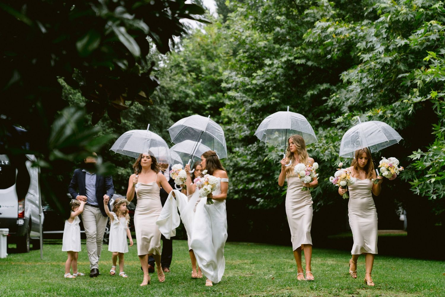 Wedding party walking with umbrellas through a lush green garden path.