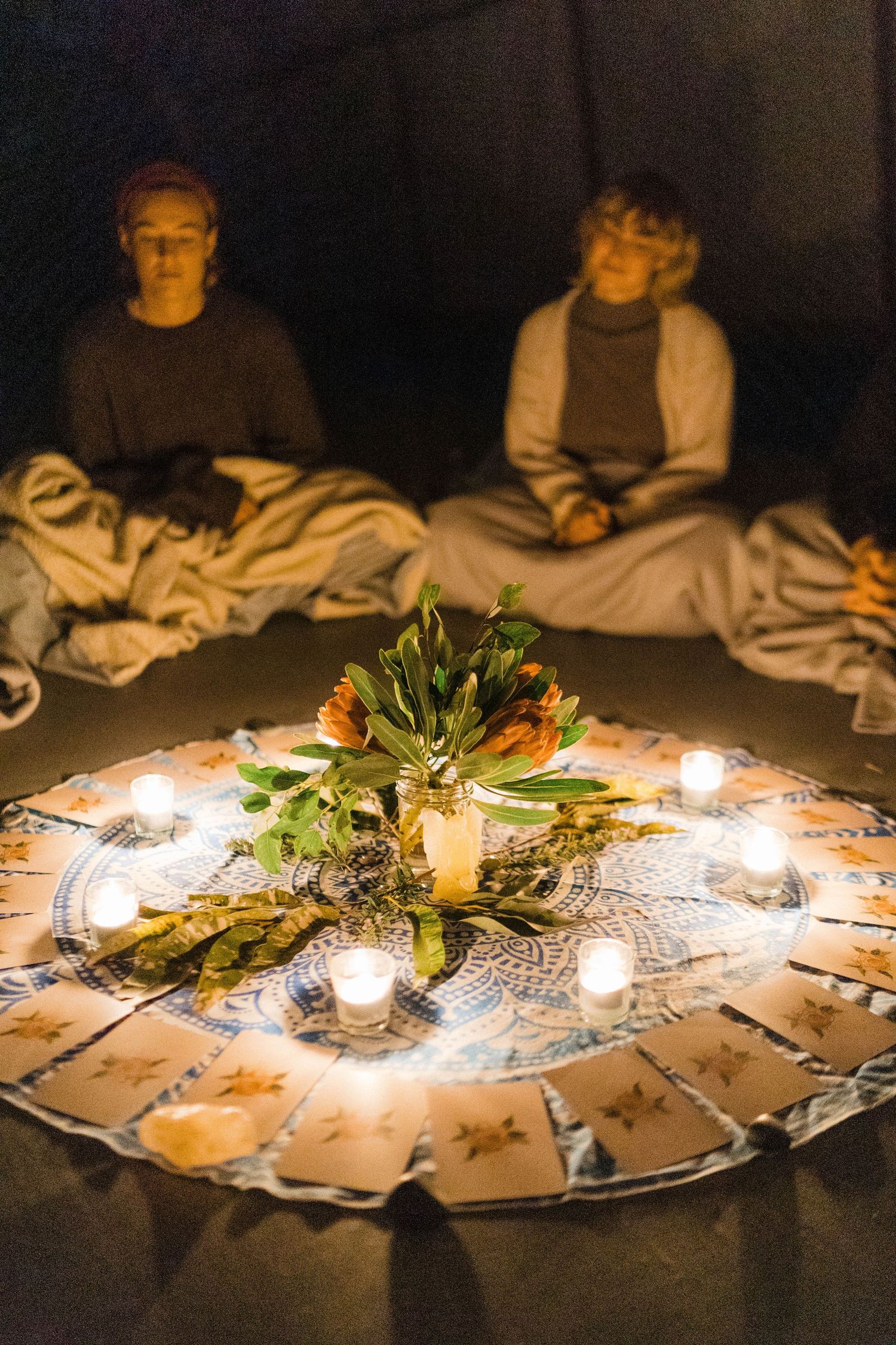 Candlelit flower arrangement on circular mandala with people sitting in darkened room.