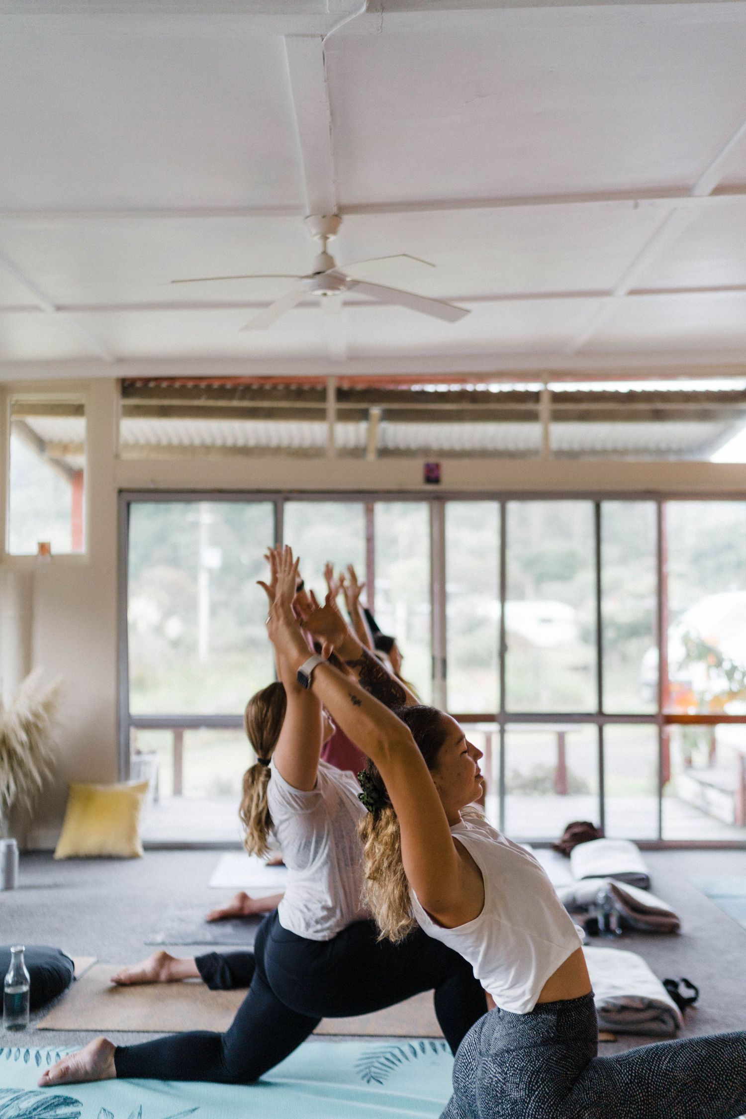 Yoga students practicing warrior pose in a bright studio space with large windows.