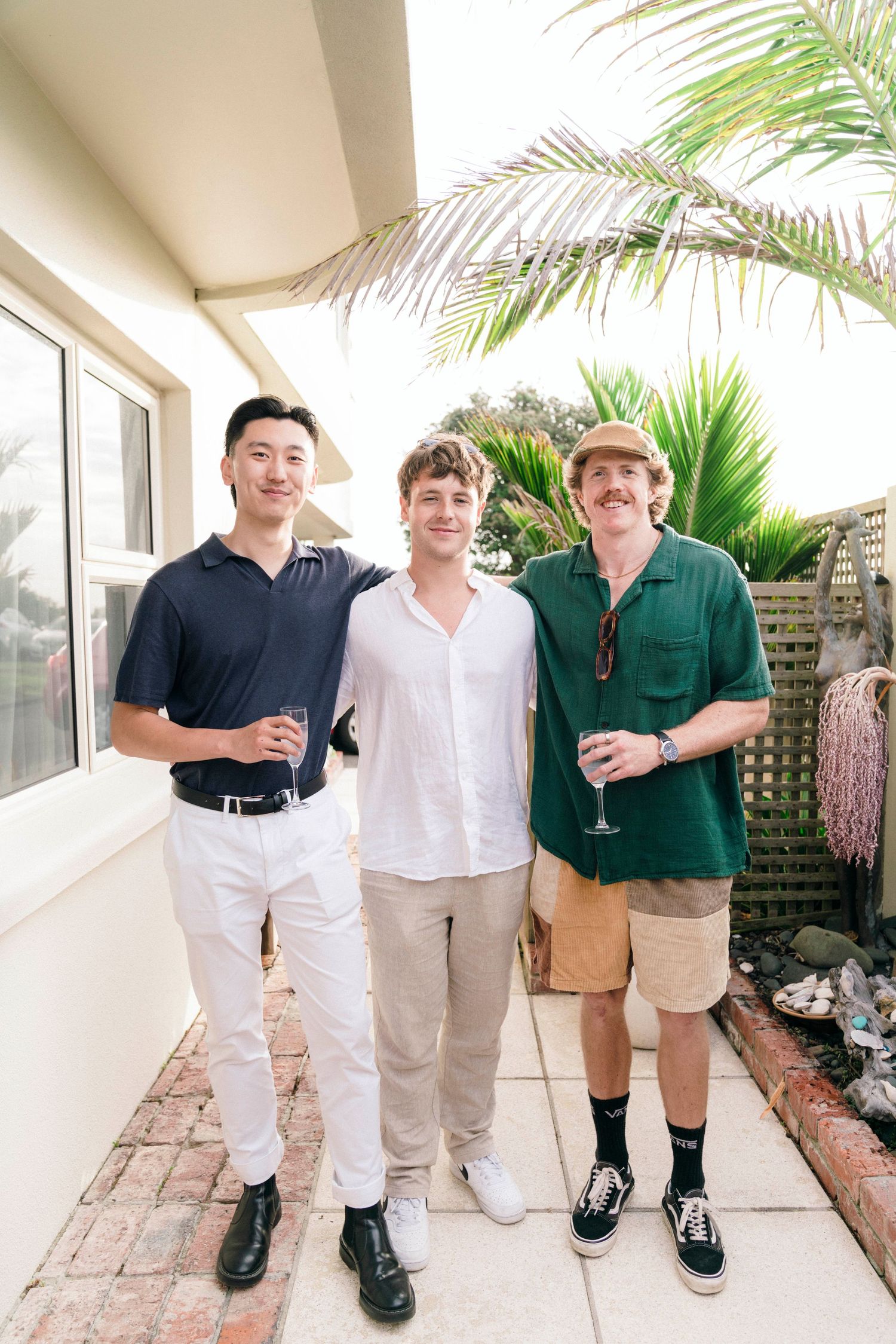 Three friends in casual attire stand together on a patio with palm trees.