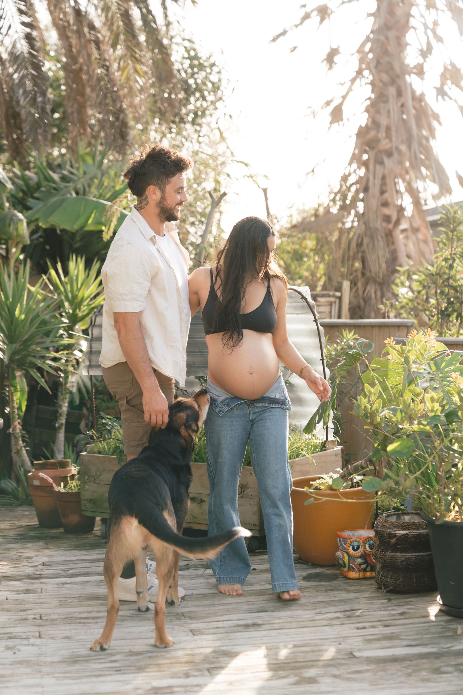 A couple and their German Shepherd dog interact playfully on a sunny patio surrounded by potted plants.