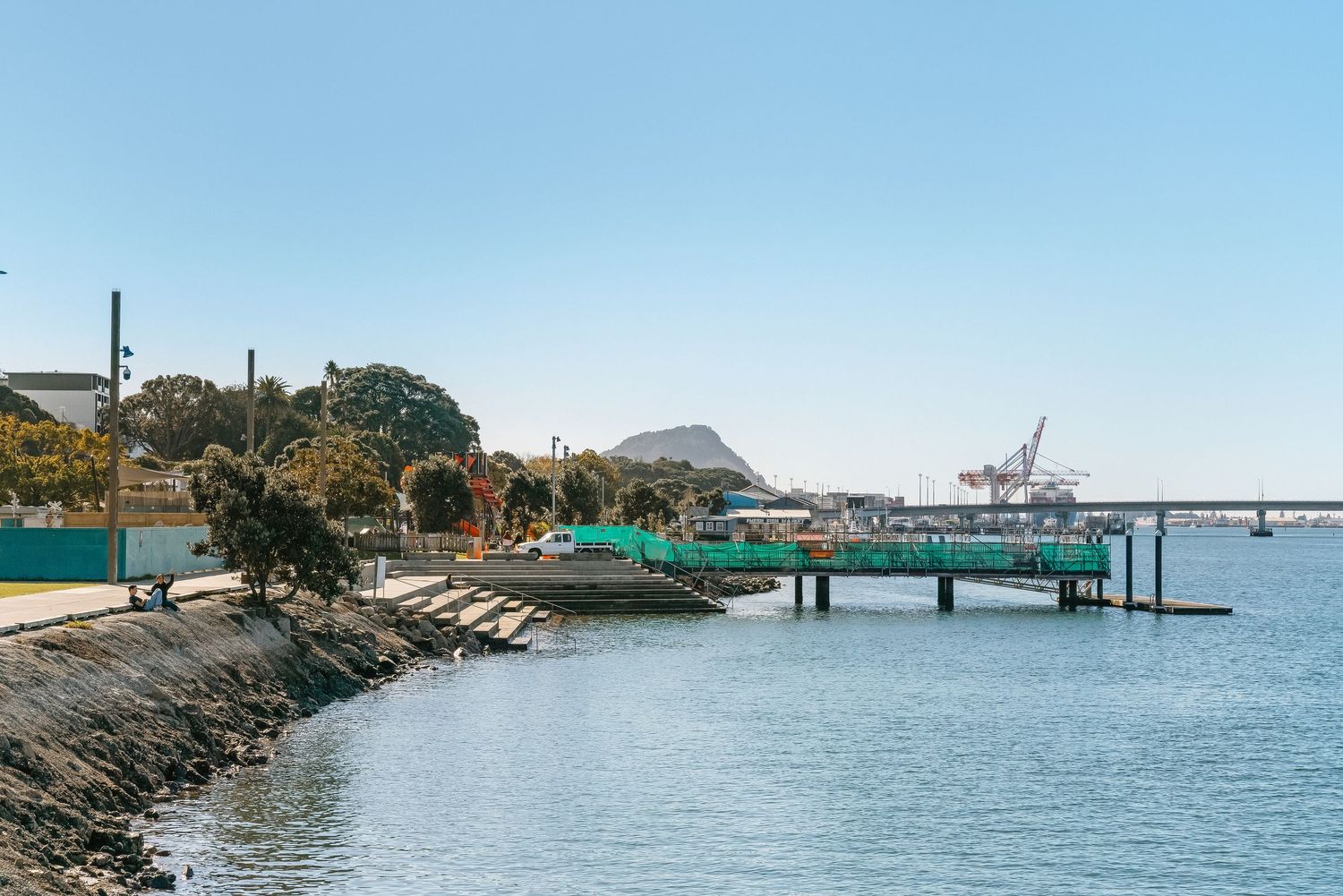 Panoramic view of a seaside marina with boats docked along green piers and mountains in the background.