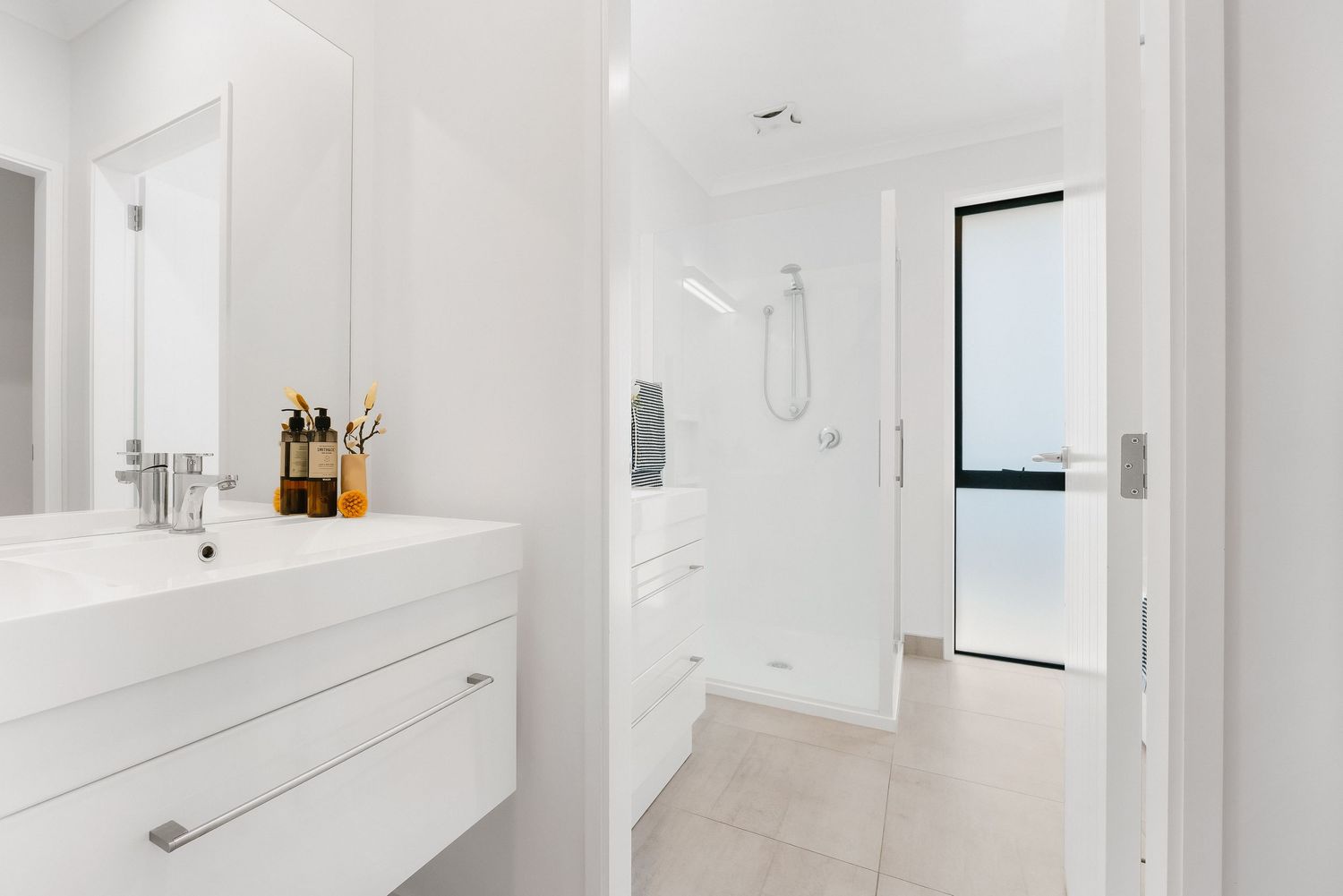 Modern white bathroom interior with glass shower enclosure, floating vanity and natural light from window.