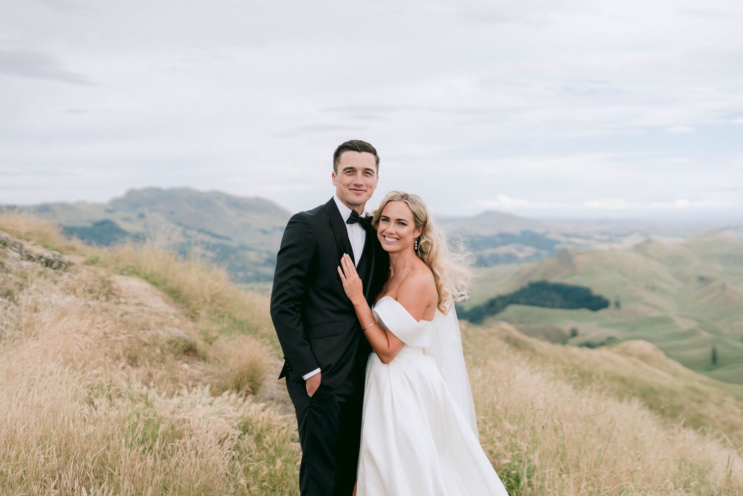 A wedding couple poses together on a scenic hilltop with rolling mountains in the background.