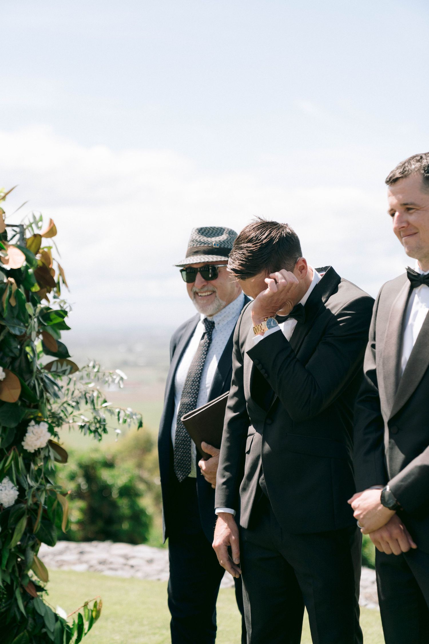 A wedding ceremony setup outdoors with suited gentlemen standing by a floral-decorated arch.