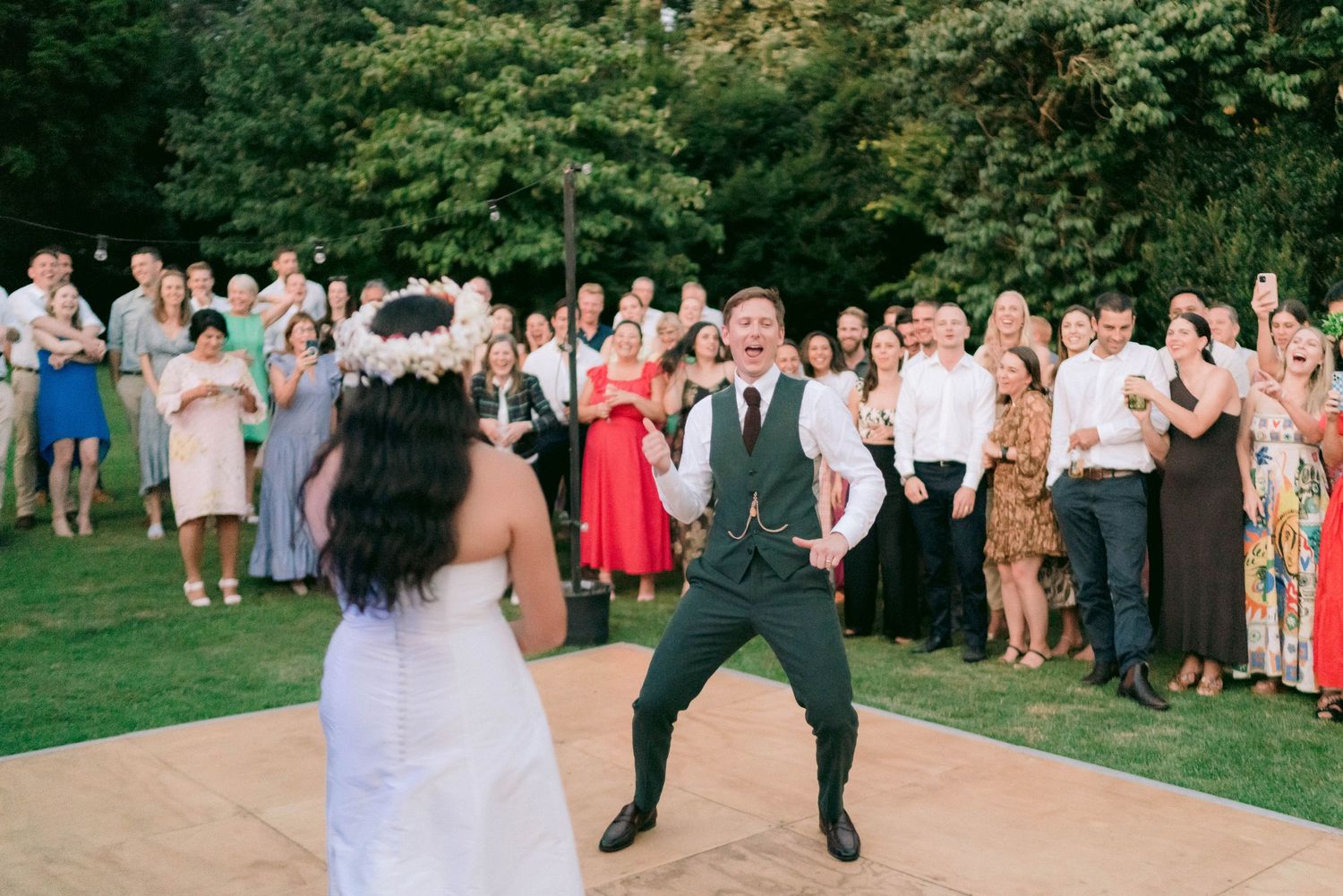 A couple dances on an outdoor dance floor while wedding guests watch and cheer from the lawn.