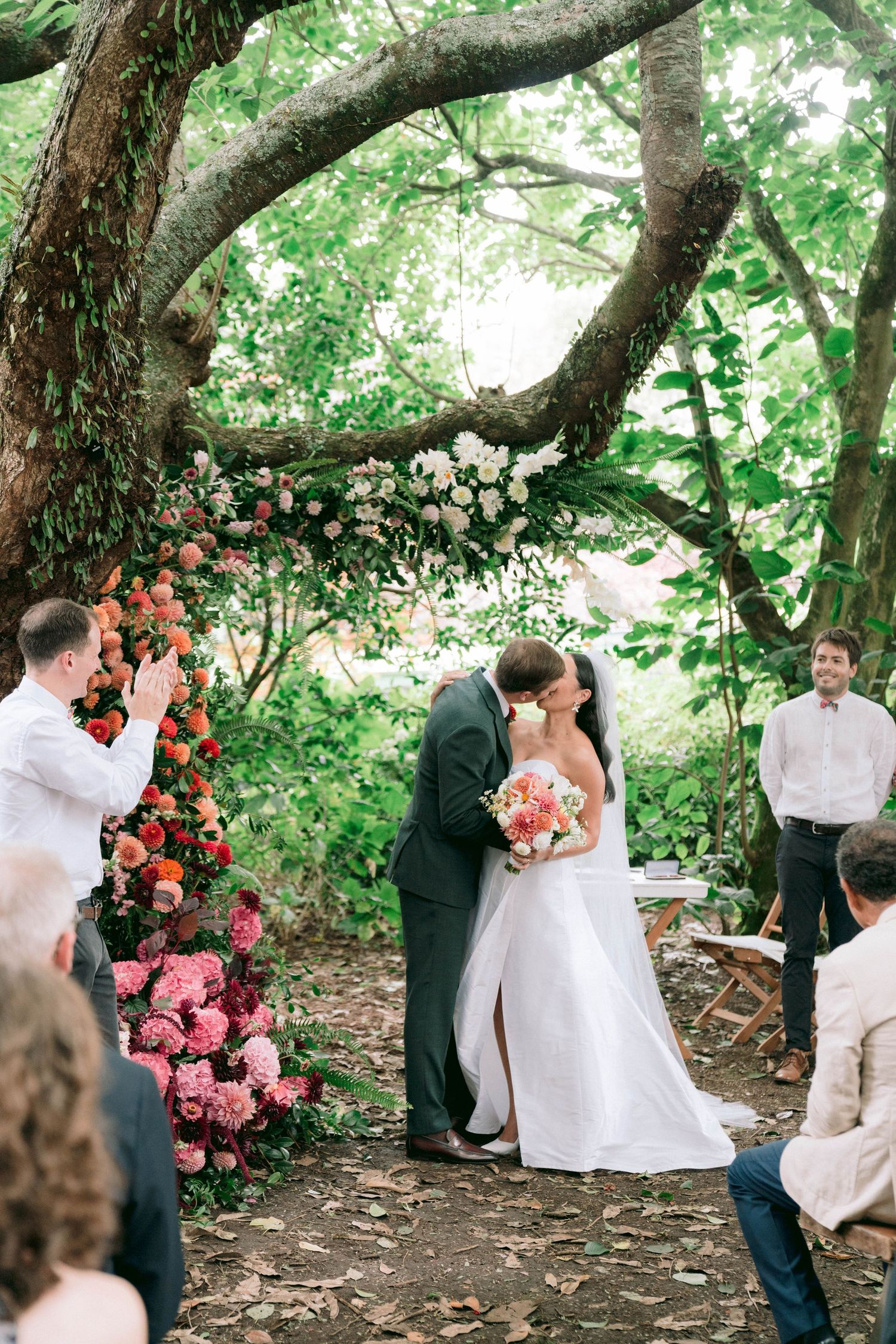 A series of outdoor wedding photos showing intimate moments beneath lush green trees with vibrant coral flowers.