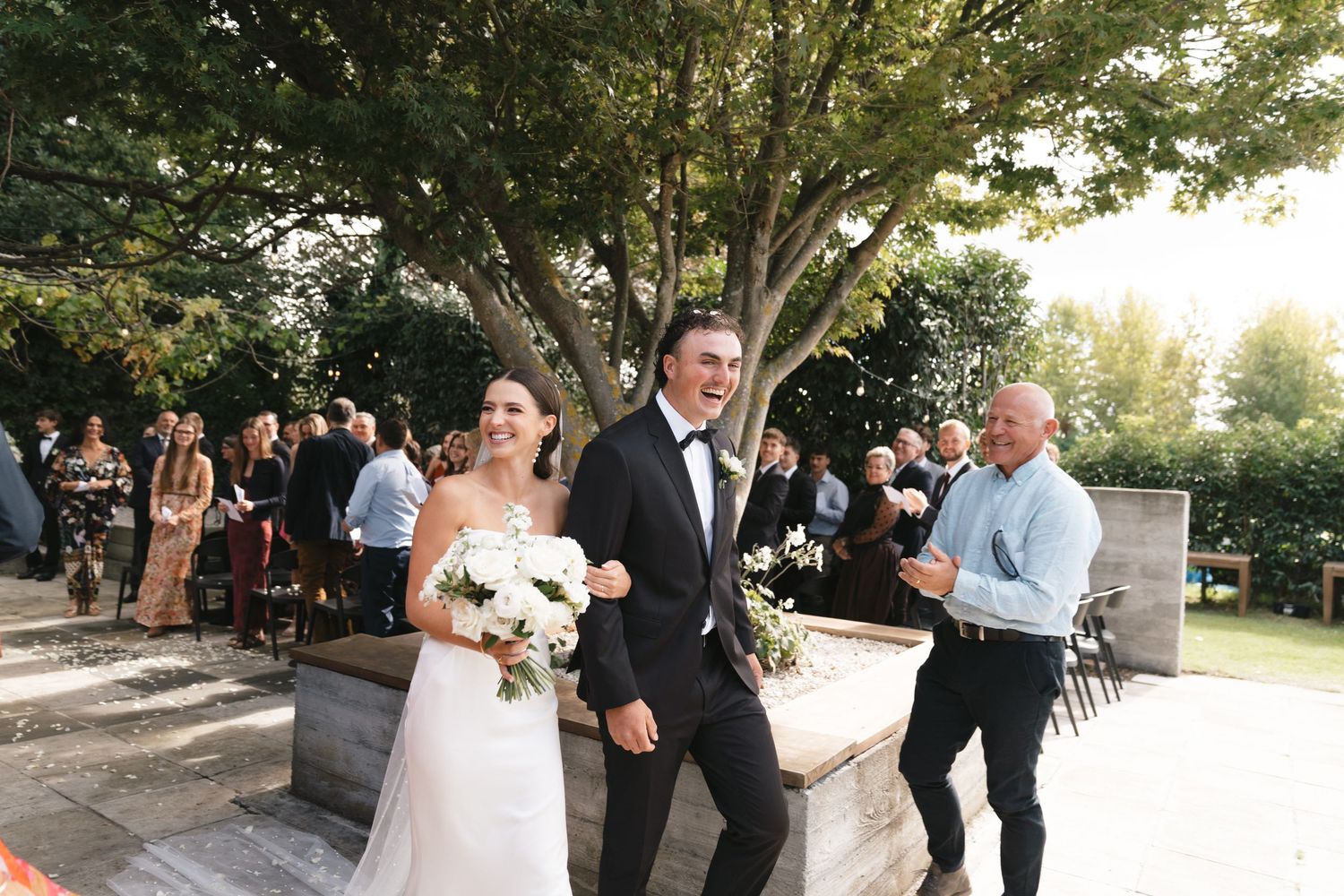 Wedding couple joyfully exiting ceremony while guests celebrate outdoors.