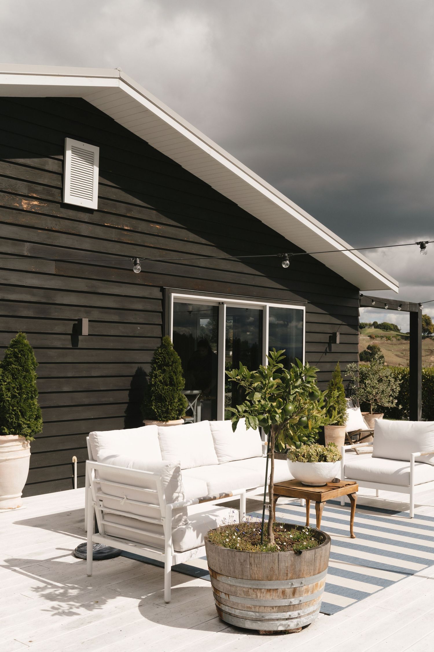 Modern outdoor patio seating area with white cushioned furniture and wooden barrel planters against dark siding.