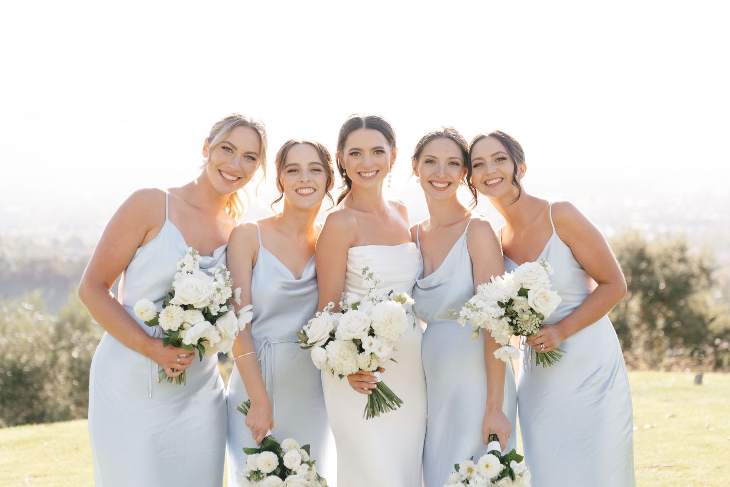 Bridesmaids in light blue dresses hold white floral bouquets while posing together outdoors on a sunny day.