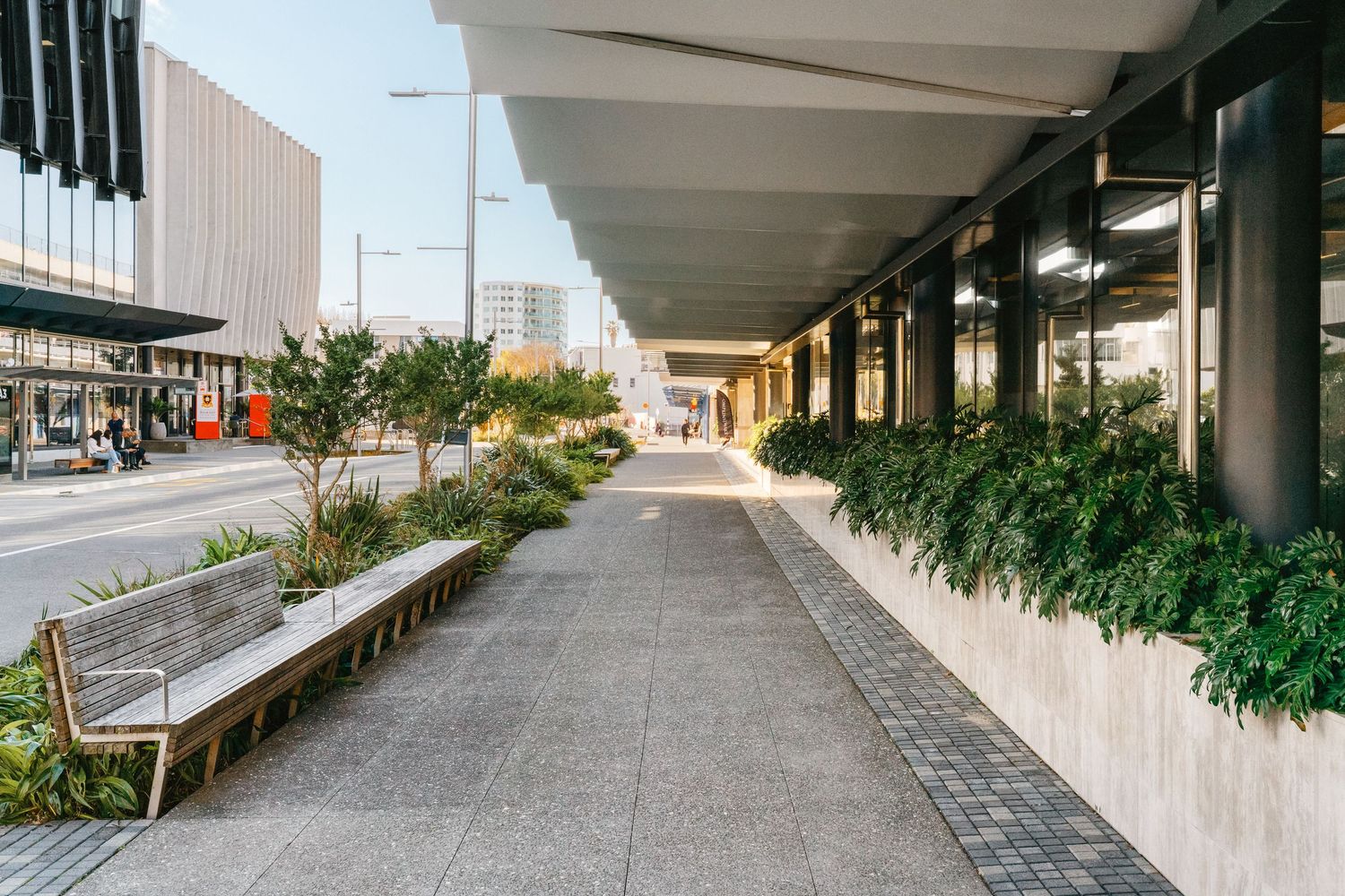 Modern urban walkway with covered colonnade, concrete benches and landscaped planters along a city street.