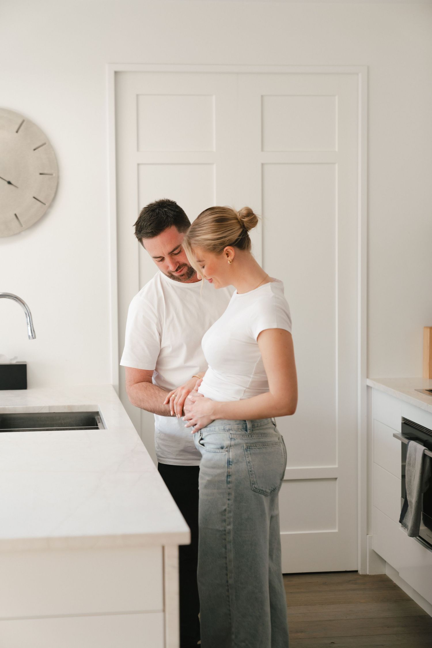 Couple sharing intimate moment in minimal white kitchen with clock on wall.