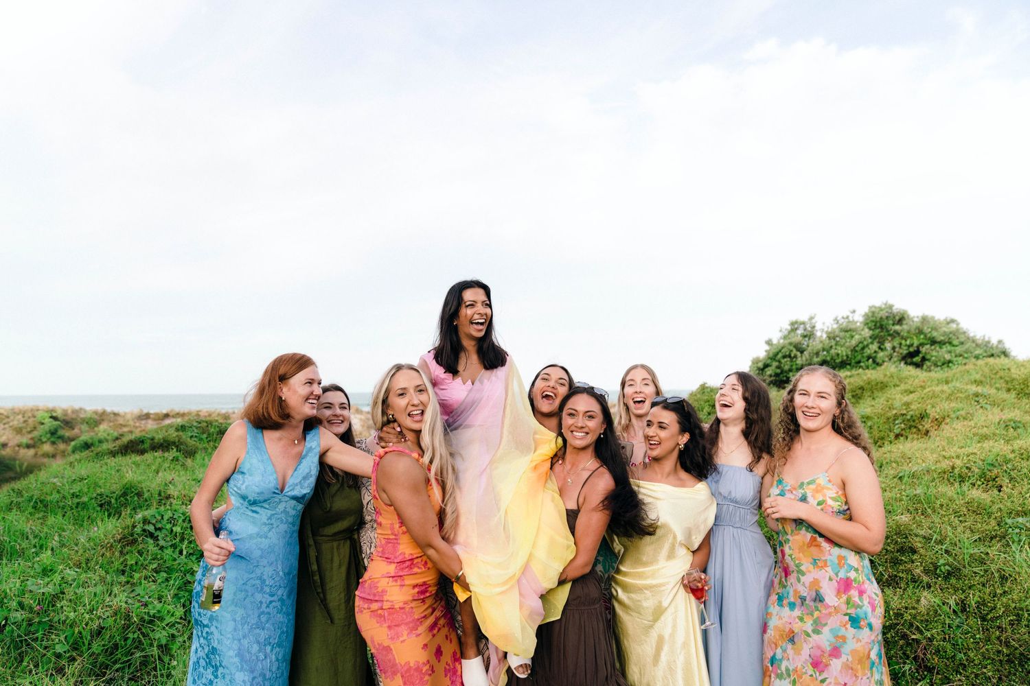 A group poses together in colorful dresses against a grassy hillside backdrop.