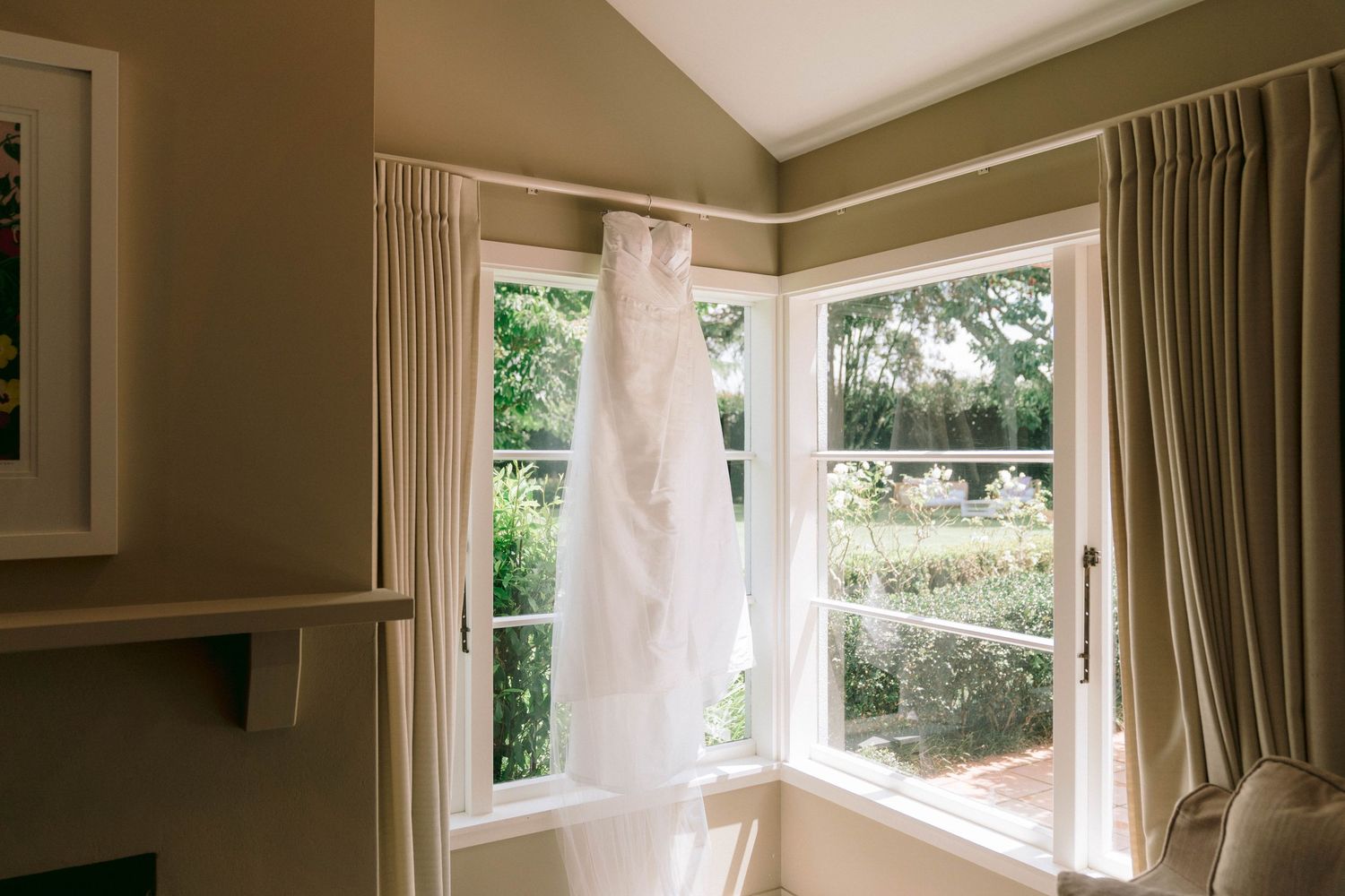 White wedding dress hanging by sunny corner windows with beige curtains.