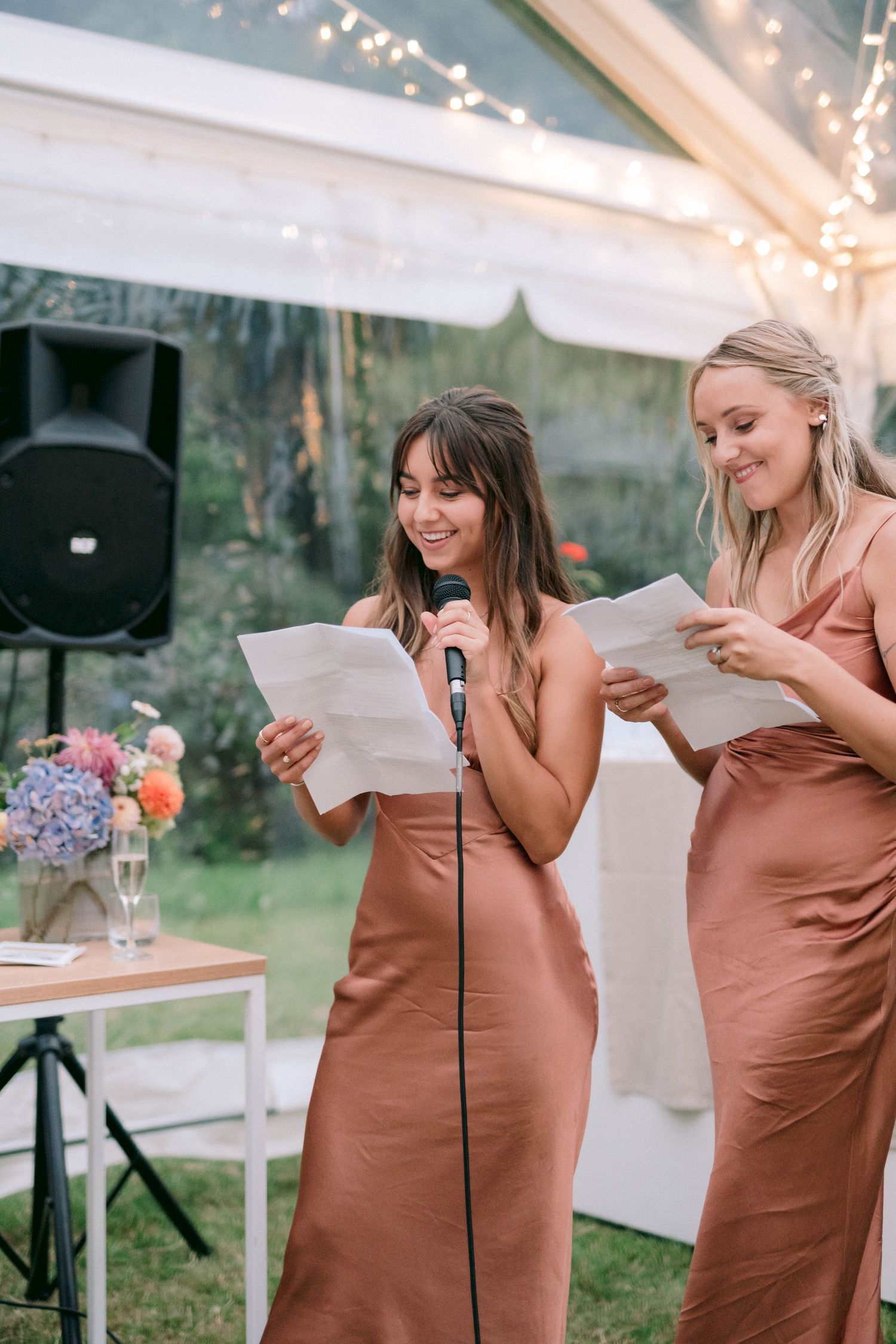 Two women in silk dresses read speeches at a wedding reception with a microphone stand.