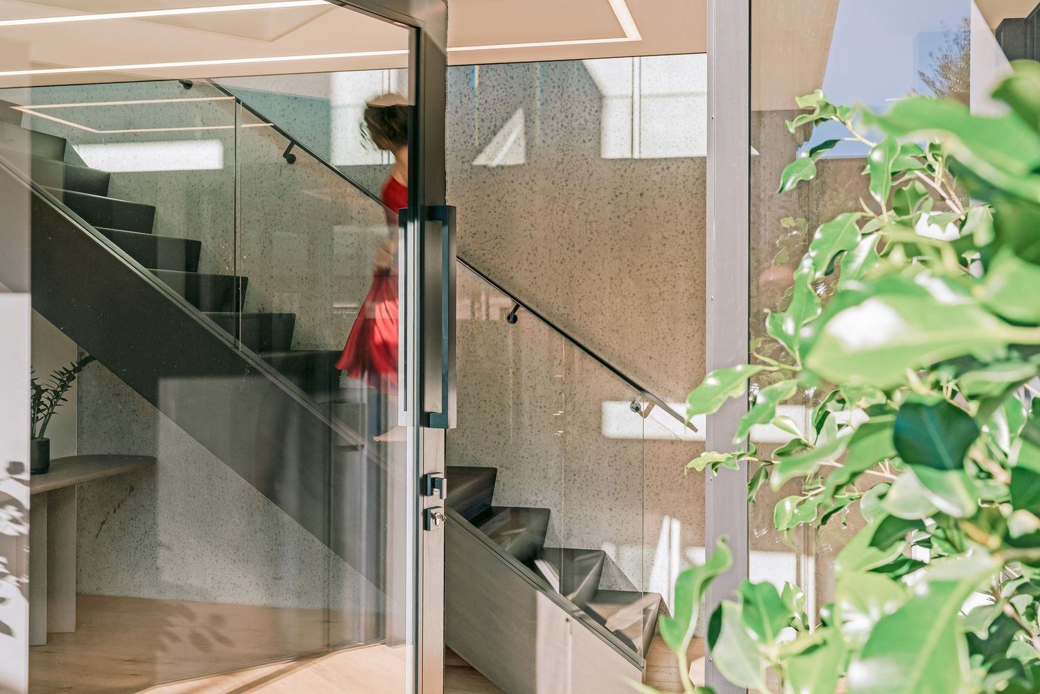 Glass staircase with black metal railings leads upwards in a minimalist white interior space.