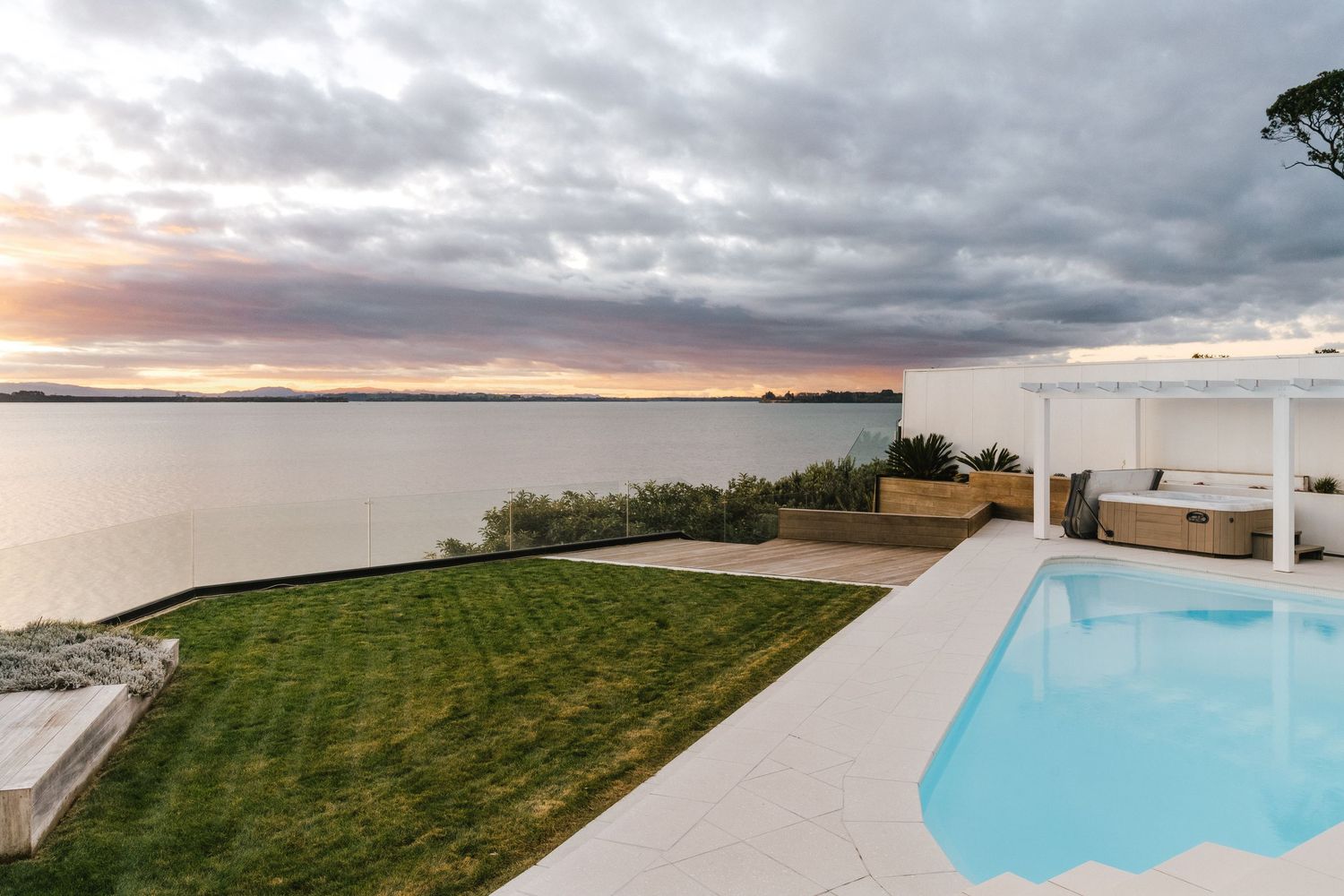 Infinity pool overlooking bay with dramatic sunset sky and manicured lawn.