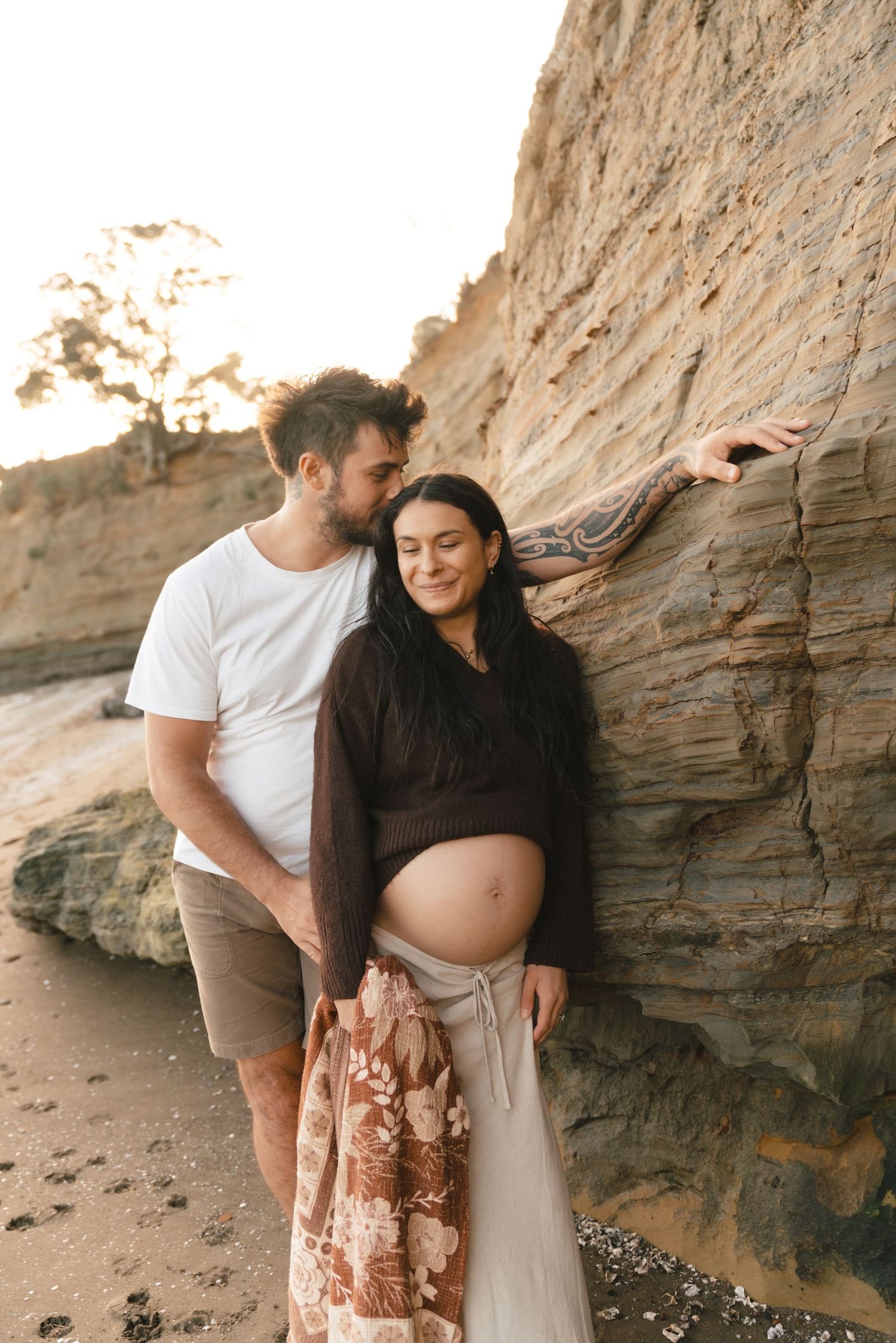 Expecting couple embraces against rocky cliffs at sunset during maternity photoshoot on beach.