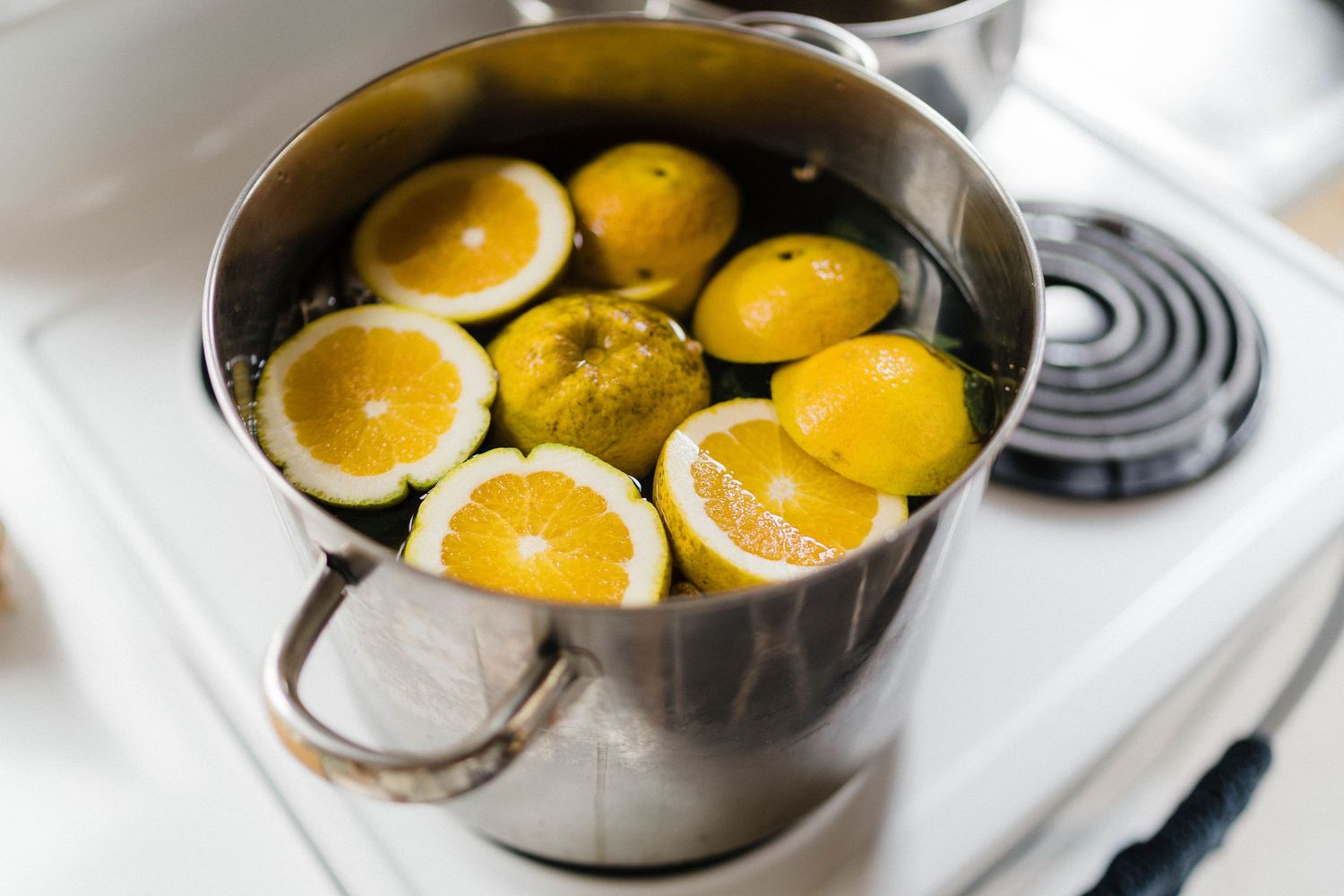 Halved oranges being simmered in a metal pot on a white stovetop.