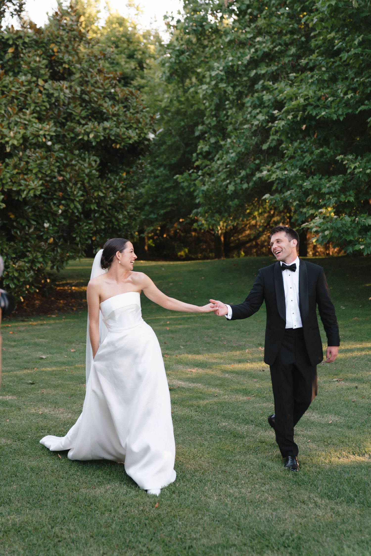 Bride and groom holding hands while walking together on green lawn during sunset.