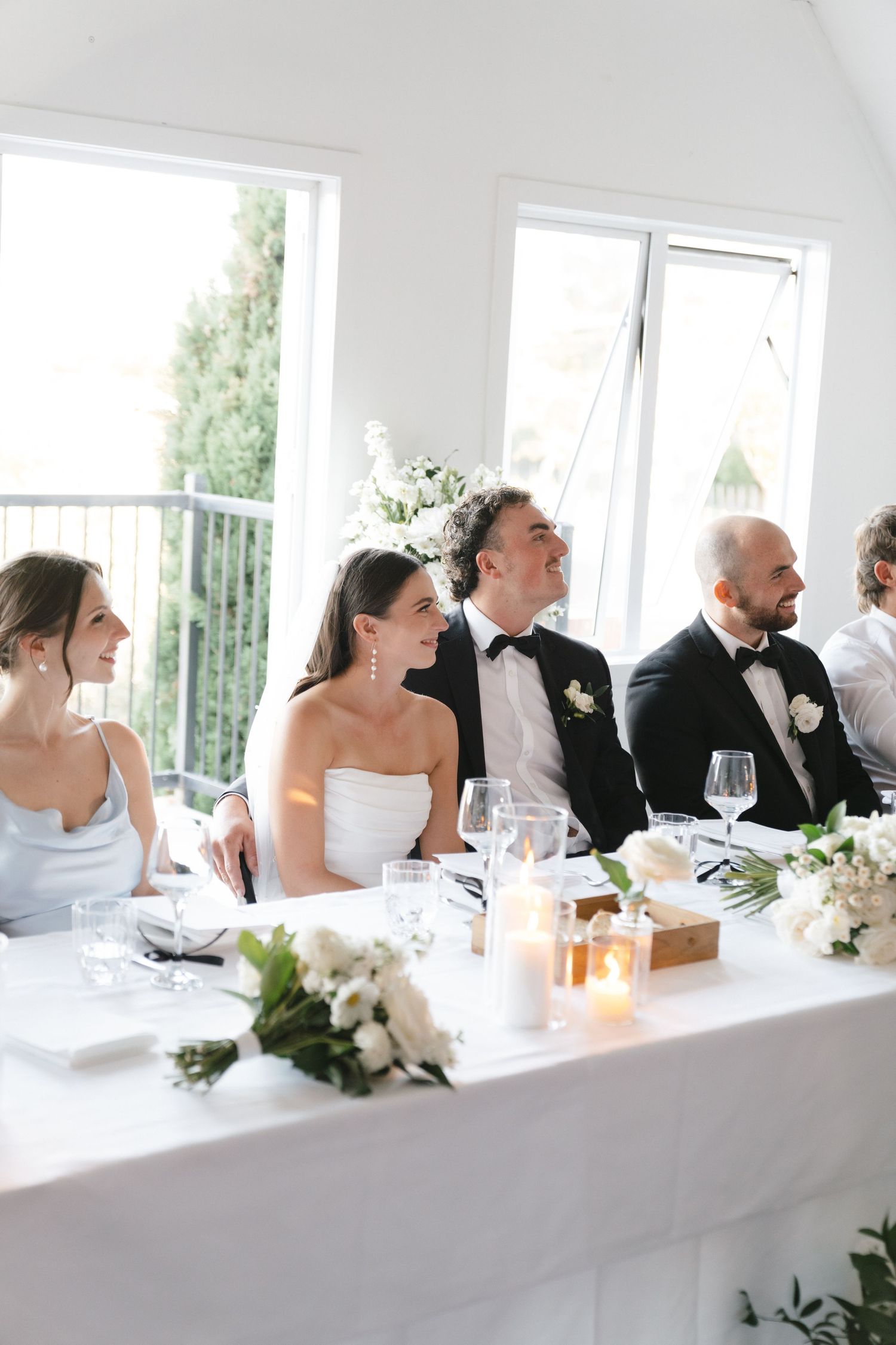 A wedding reception party seated at an elegant white-clothed head table with wine glasses and floral centerpieces.