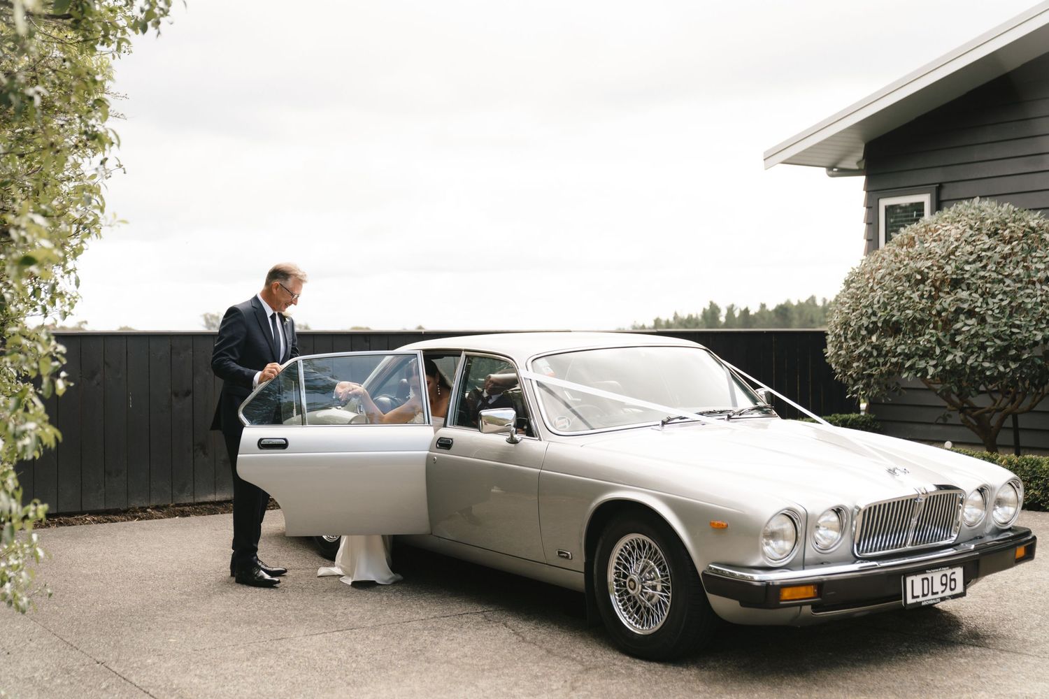 A classic silver Jaguar XJ luxury car parked outside a house with someone opening the door.