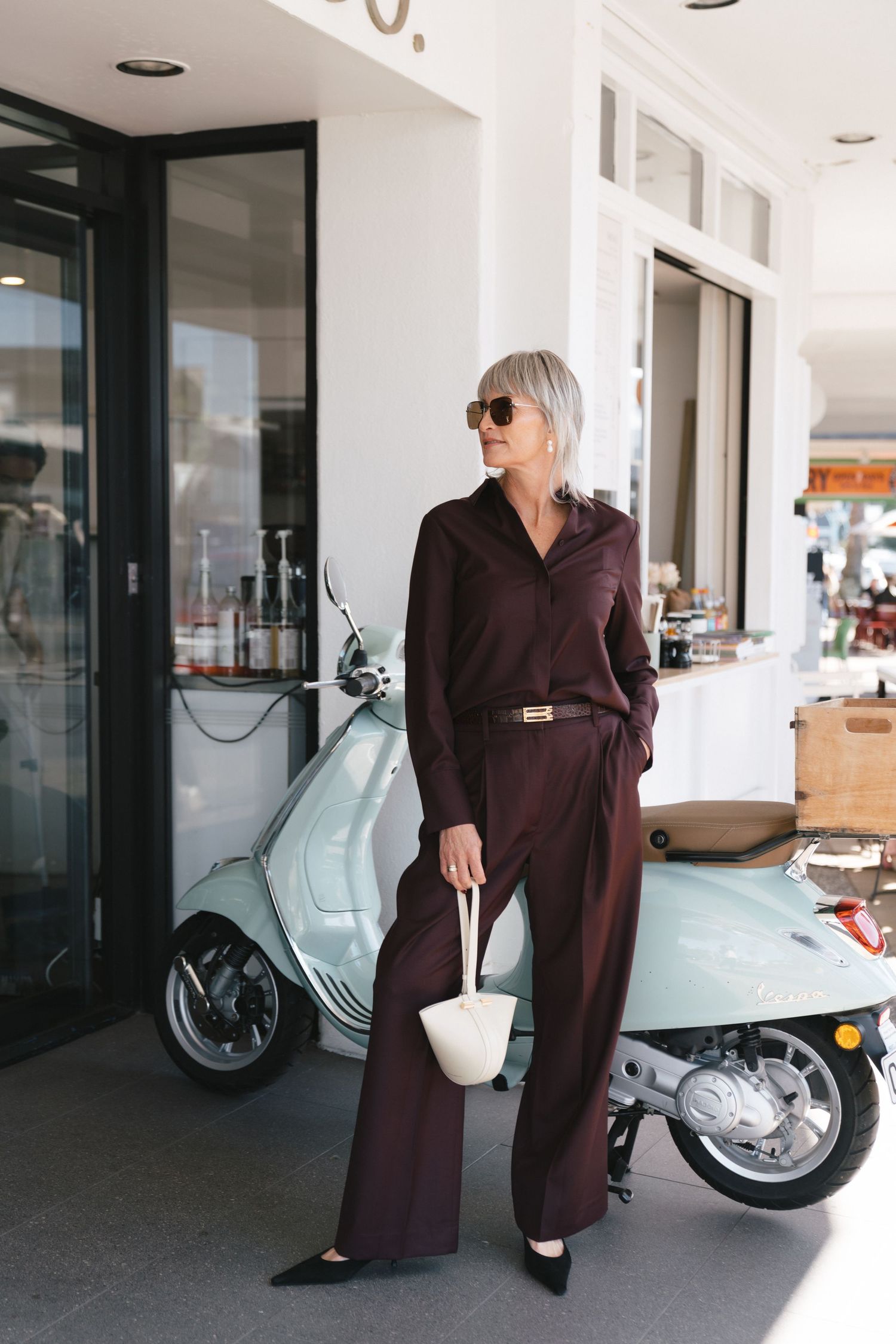 Fashionable outfit featuring a brown jumpsuit and white handbag, posed next to a light blue vintage Vespa scooter.