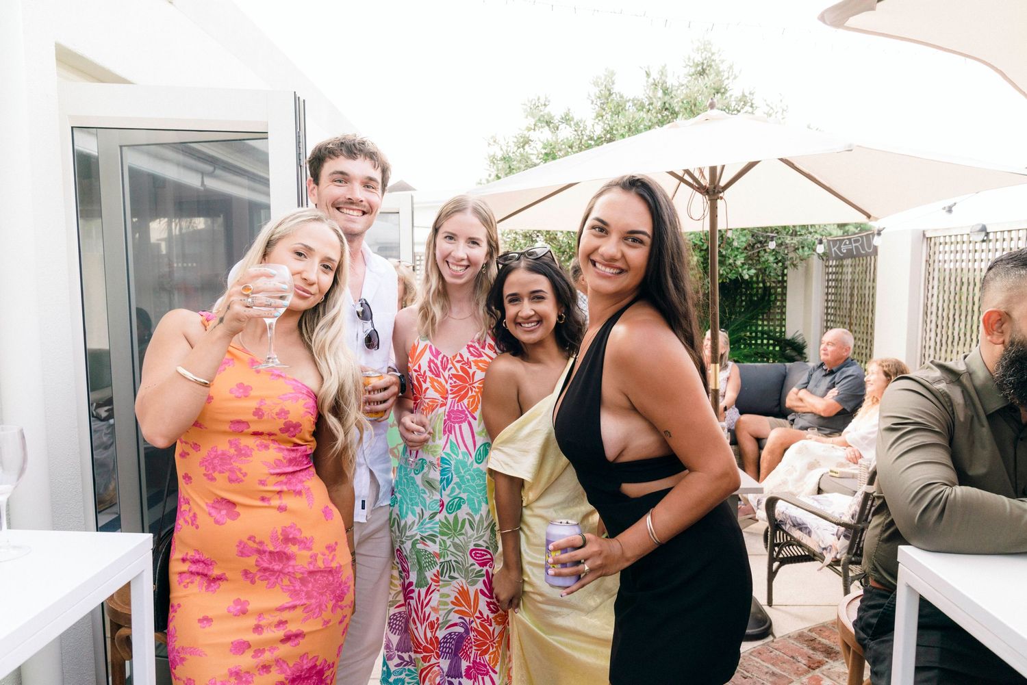 A group celebrates outdoors on a patio with bright floral dresses and joyful expressions.