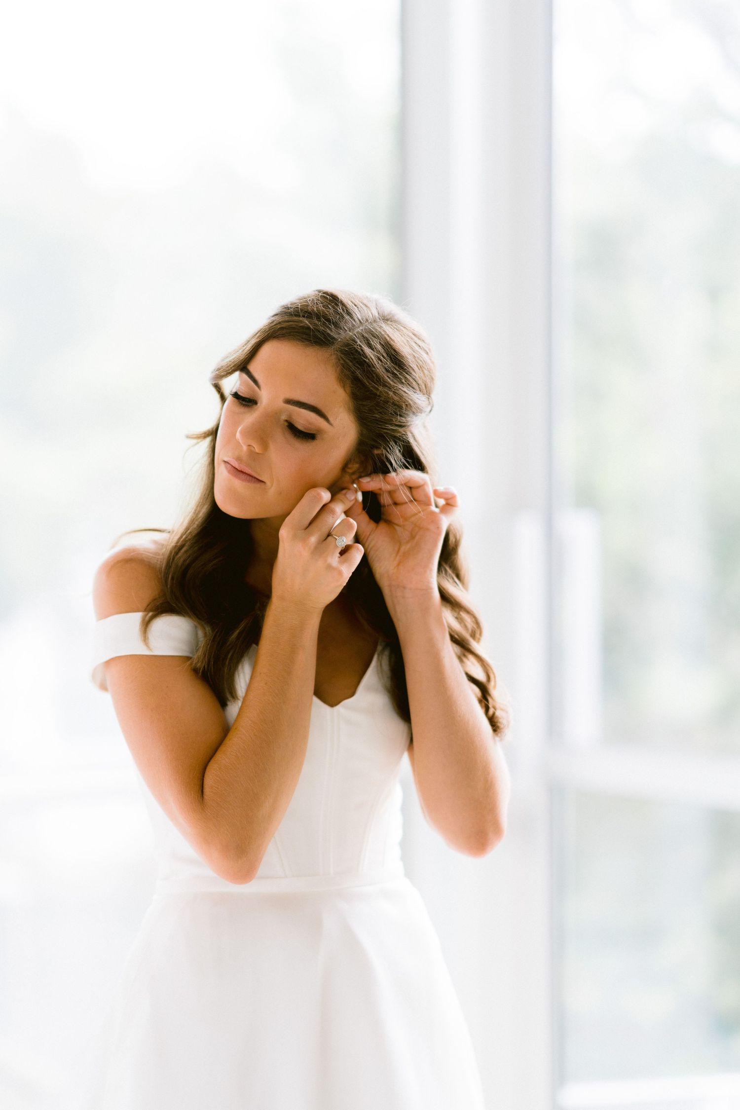 A bride getting ready on her wedding day in a bright sunlit room wearing a white dress.