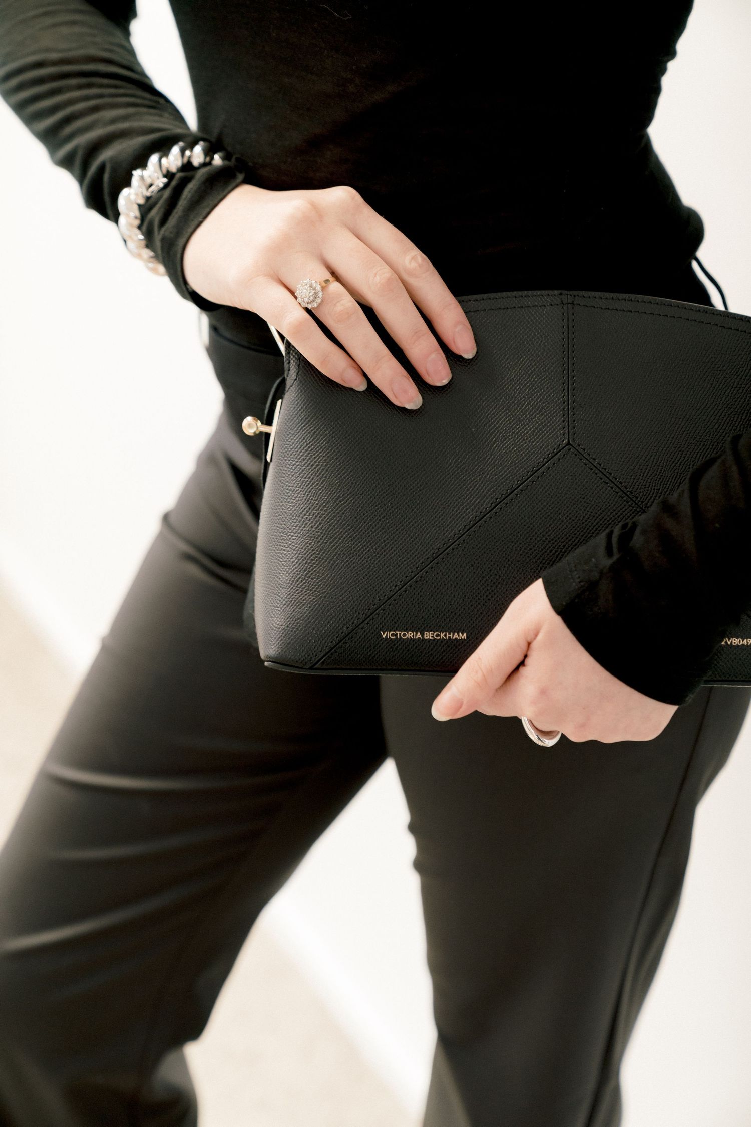 Hands holding a sleek black leather Michael Kors clutch against black leather pants creating a monochromatic look.