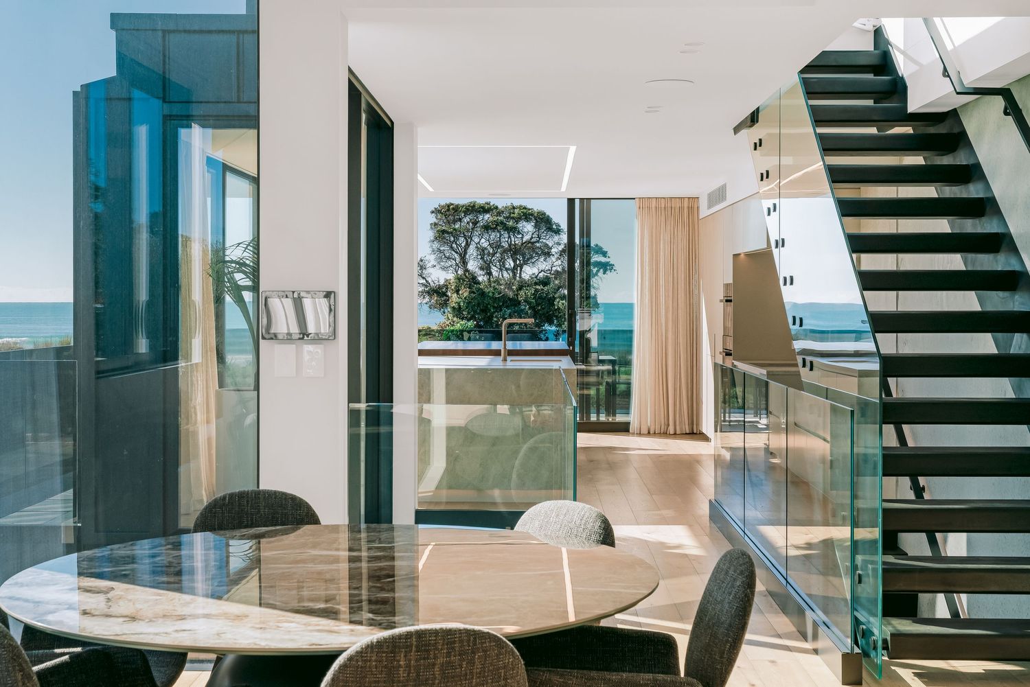 Modern coastal apartment interior with glass balcony, round dining table and floating staircase overlooking ocean views.
