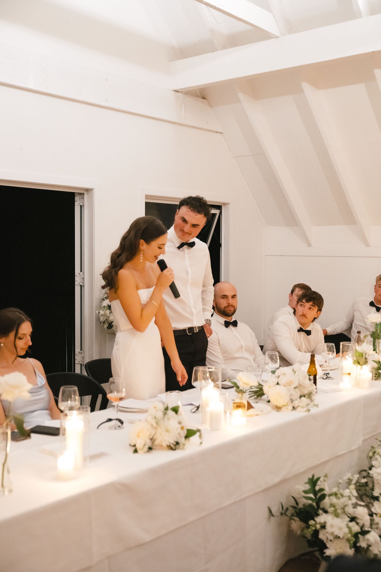 Elegant wedding reception dinner with guests seated at a long white table decorated with flowers and candles.