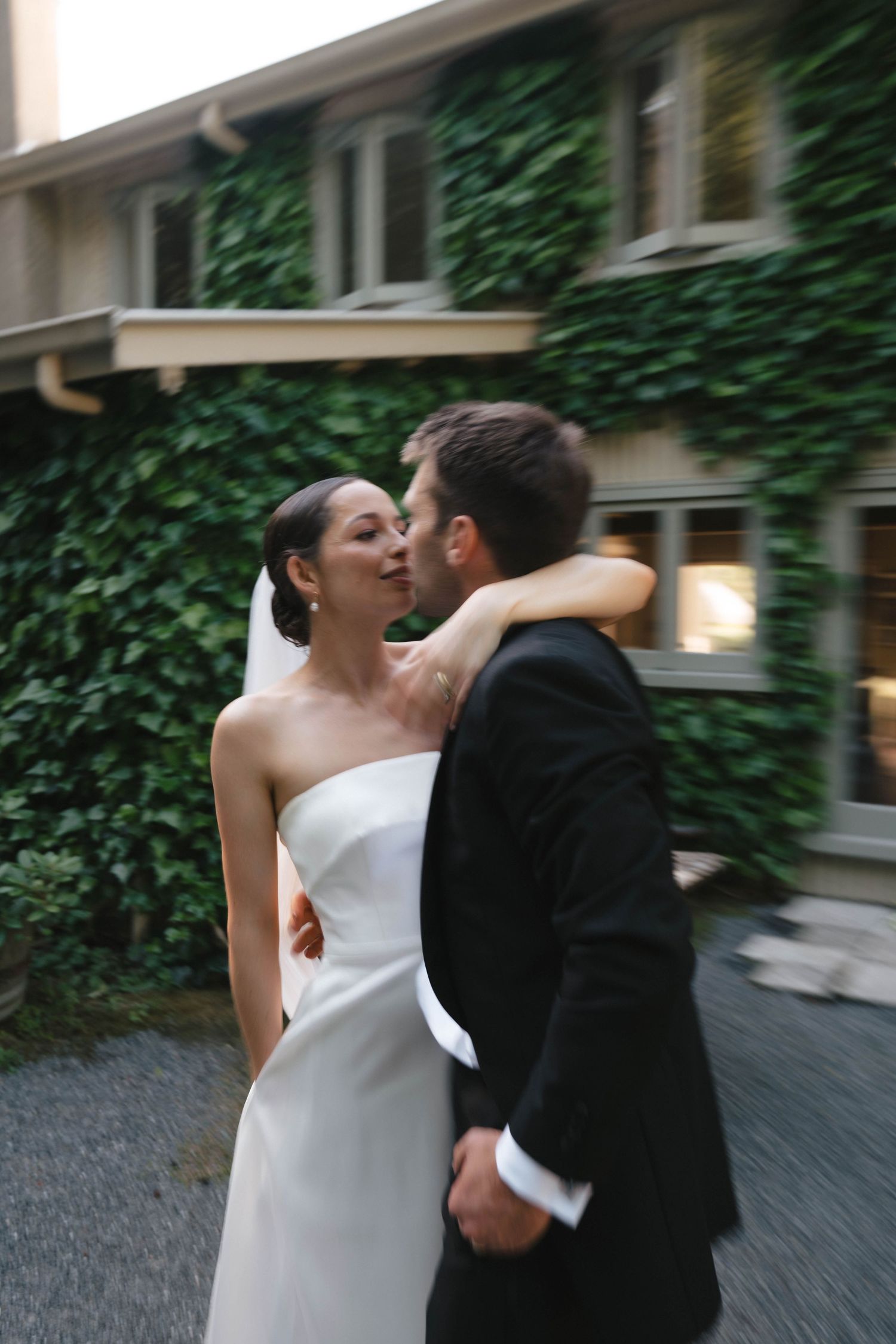 A joyful couple in wedding attire embrace and laugh together outside a building with ivy-covered walls.
