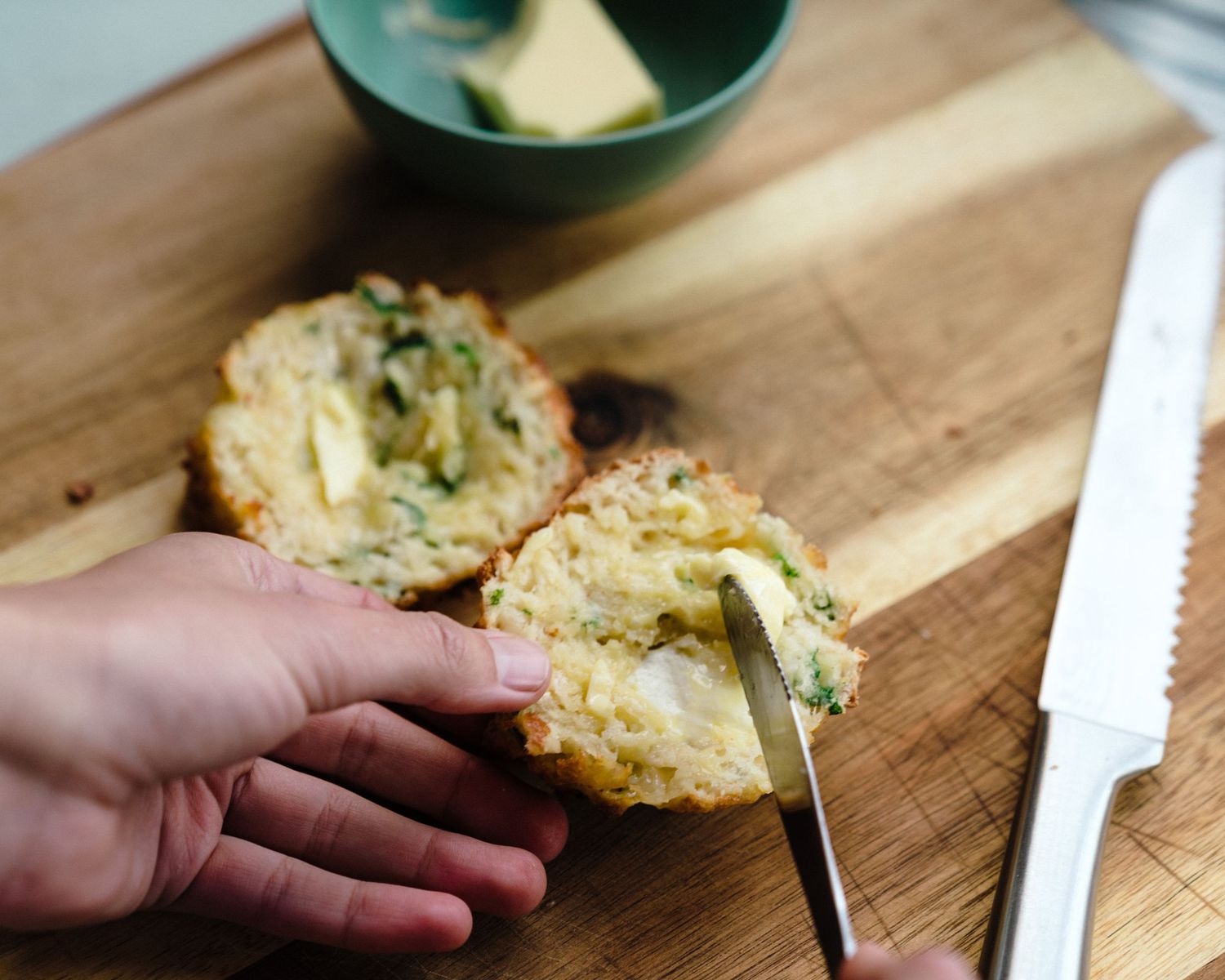 Hands slicing warm homemade cookies on a wooden cutting board with a serrated knife.