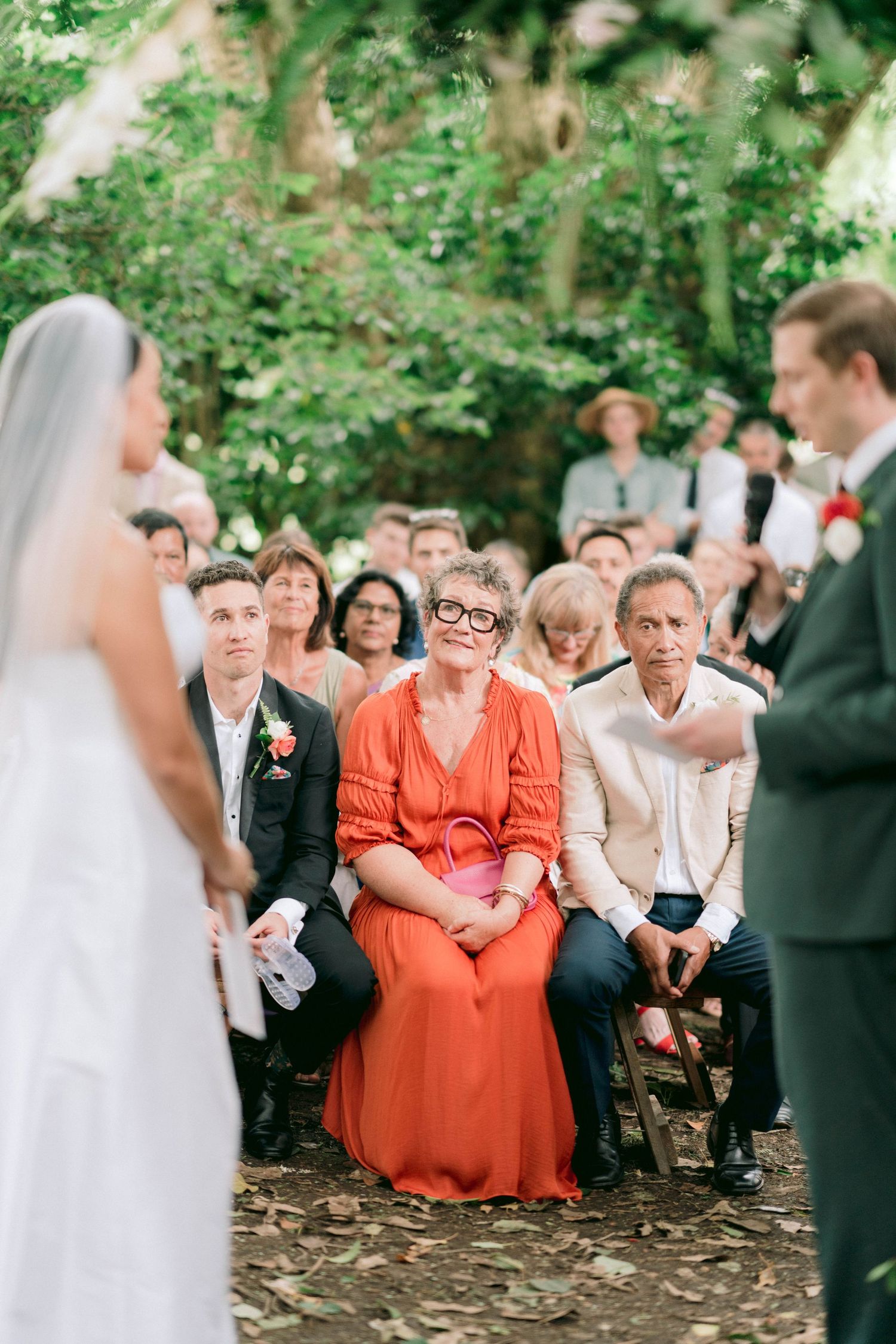 Outdoor wedding ceremony with guests seated on wooden chairs beneath trees.