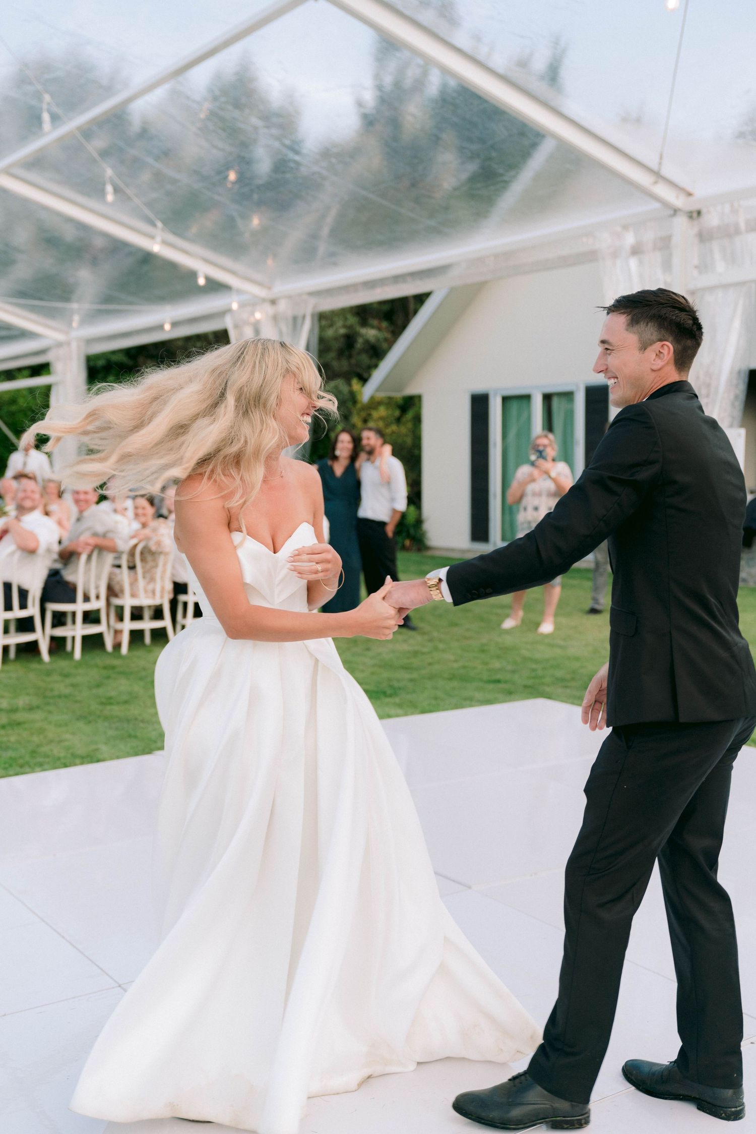 A wedding couple sharing their first dance under a clear tent with guests watching from white chairs.