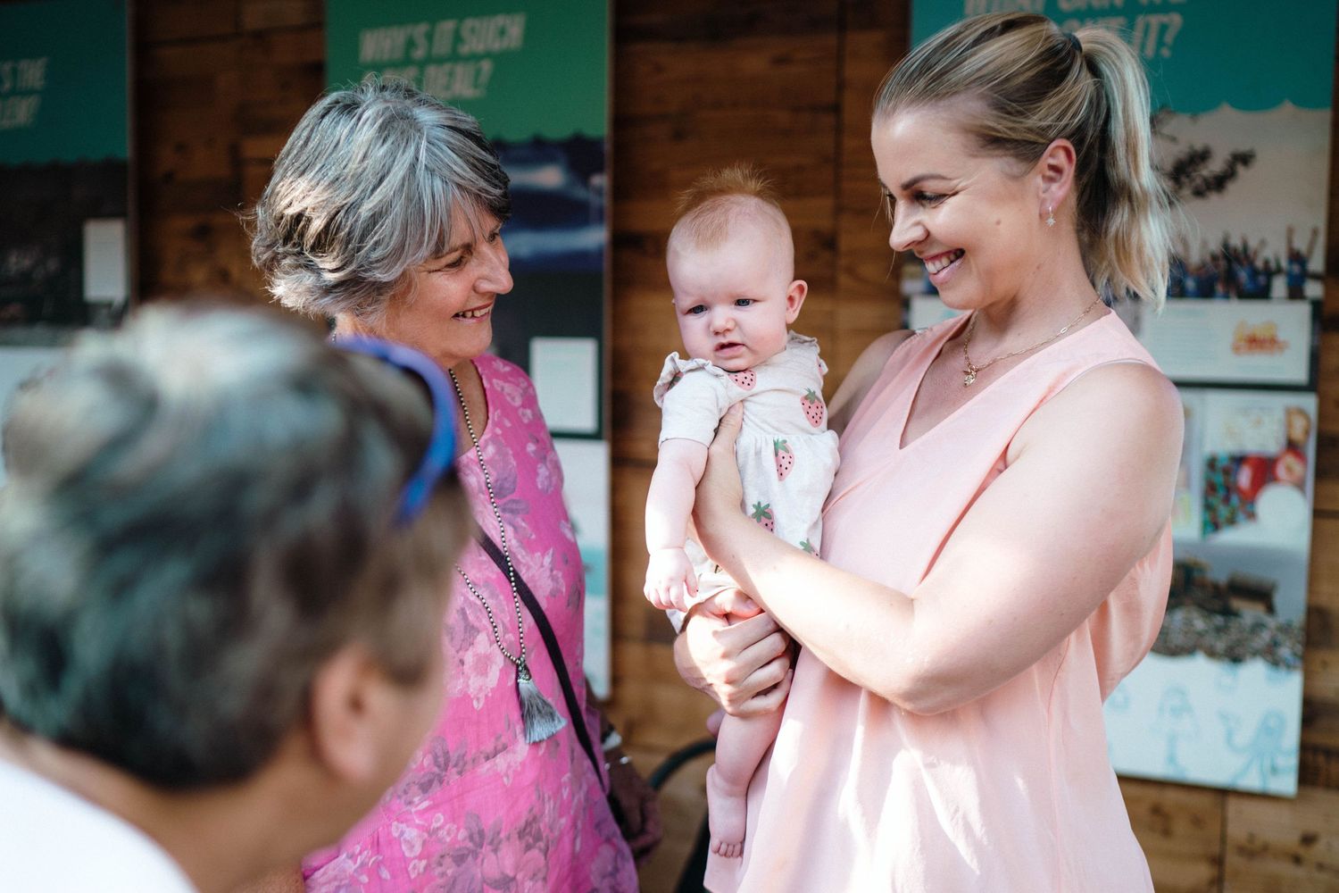Smiling people wearing pink outfits gather together on a sunny day outside a building with a young infant.