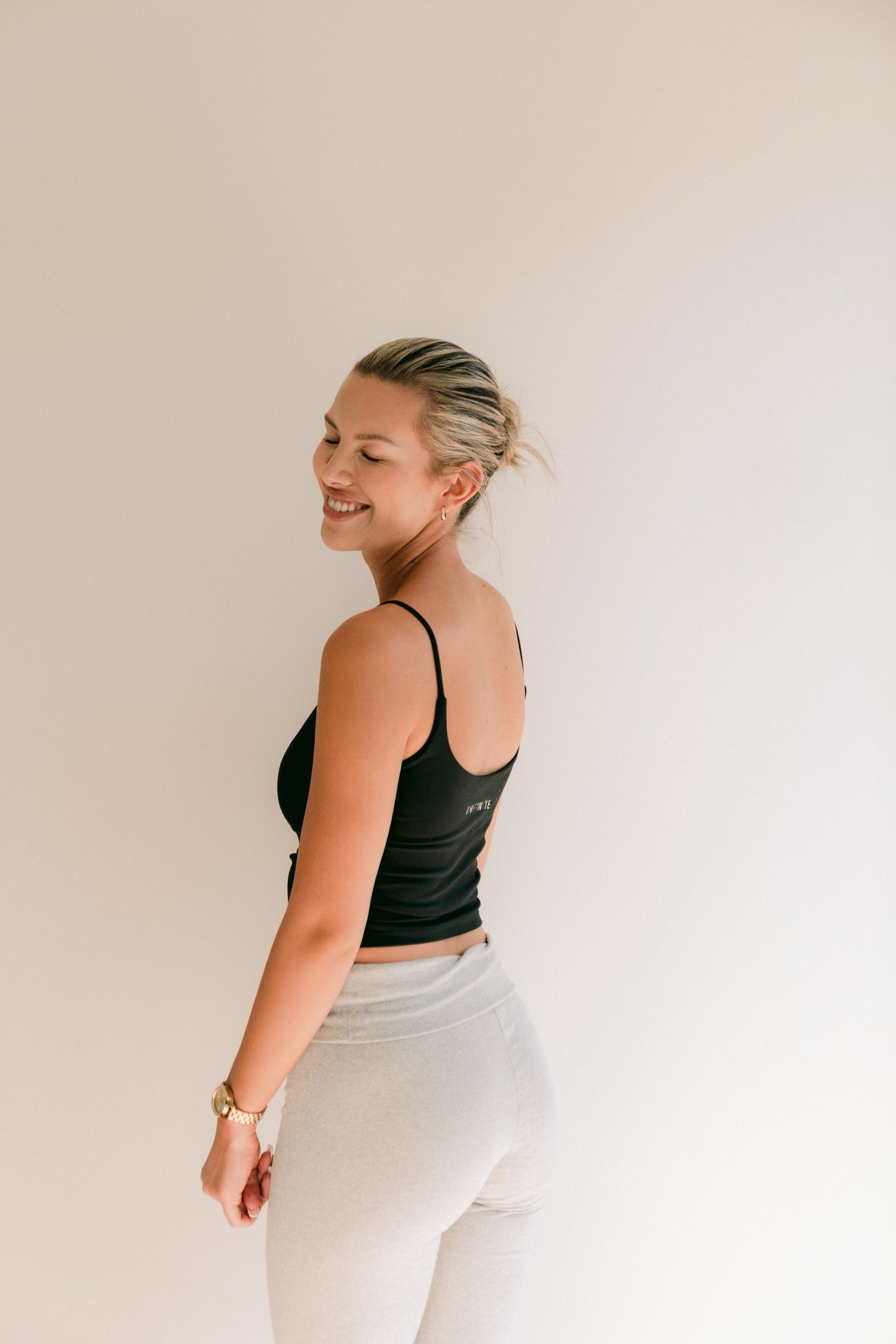 Model in black cami and white pants turns to side with natural smile against minimalist background.